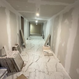 MjHorizonLine Sp Z o.o. - Długi korytarz w trakcie remontu, z marmurową podłogą, ułożoną częściowo z płytek, z widocznymi workami z zaprawą, płytkami i narzędziami glazurniczymi.