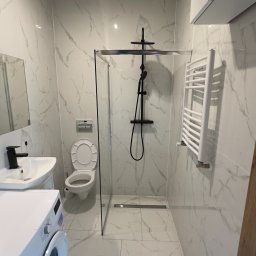 MjHorizonLine Sp Z o.o. - Biała łazienka z marmurowymi płytkami, kabiną prysznicową typu walk-in z czarną armaturą, toaletą, umywalką z czarną baterią i pralką Sharp.