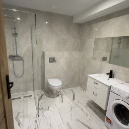MjHorizonLine Sp Z o.o. - Jasna łazienka po remoncie z kabiną prysznicową typu walk-in, toaletą podwieszaną, białą szafką z umywalką i pralką, wykończona płytkami imitującymi marmur.