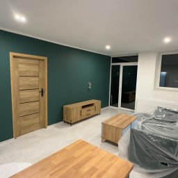 MjHorizonLine Sp Z o.o. - Wnętrze pokoju po malowaniu ścian na ciemnozielono, z jasnymi drewnianymi meblami, drzwiami i podłogą, częściowo zabezpieczonym folią meblem i widocznymi instalacjami elektrycznymi na ścianie.