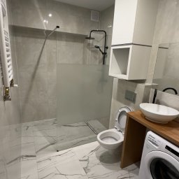 MjHorizonLine Sp Z o.o. - Nowoczesna łazienka z kabiną prysznicową typu walk-in, umywalką nablatową na drewnianym blacie, wiszącą szafką i pralką Candy, wykończona płytkami imitującymi marmur.
