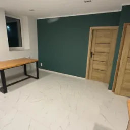 Pomalowane na ciemnozielono wnętrze pokoju z dwiema drewnianymi drzwiami z poziomymi wstawkami, marmurową podłogą i dwoma drewnianymi stołami z czarnymi nogami.