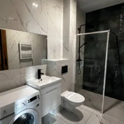 Nowoczesna łazienka w bieli i czerni z marmurowymi płytkami, kabina prysznicowa typu walk-in z czarną armaturą, biała umywalka z szafką, toaleta podwieszana i pralka Electrolux.