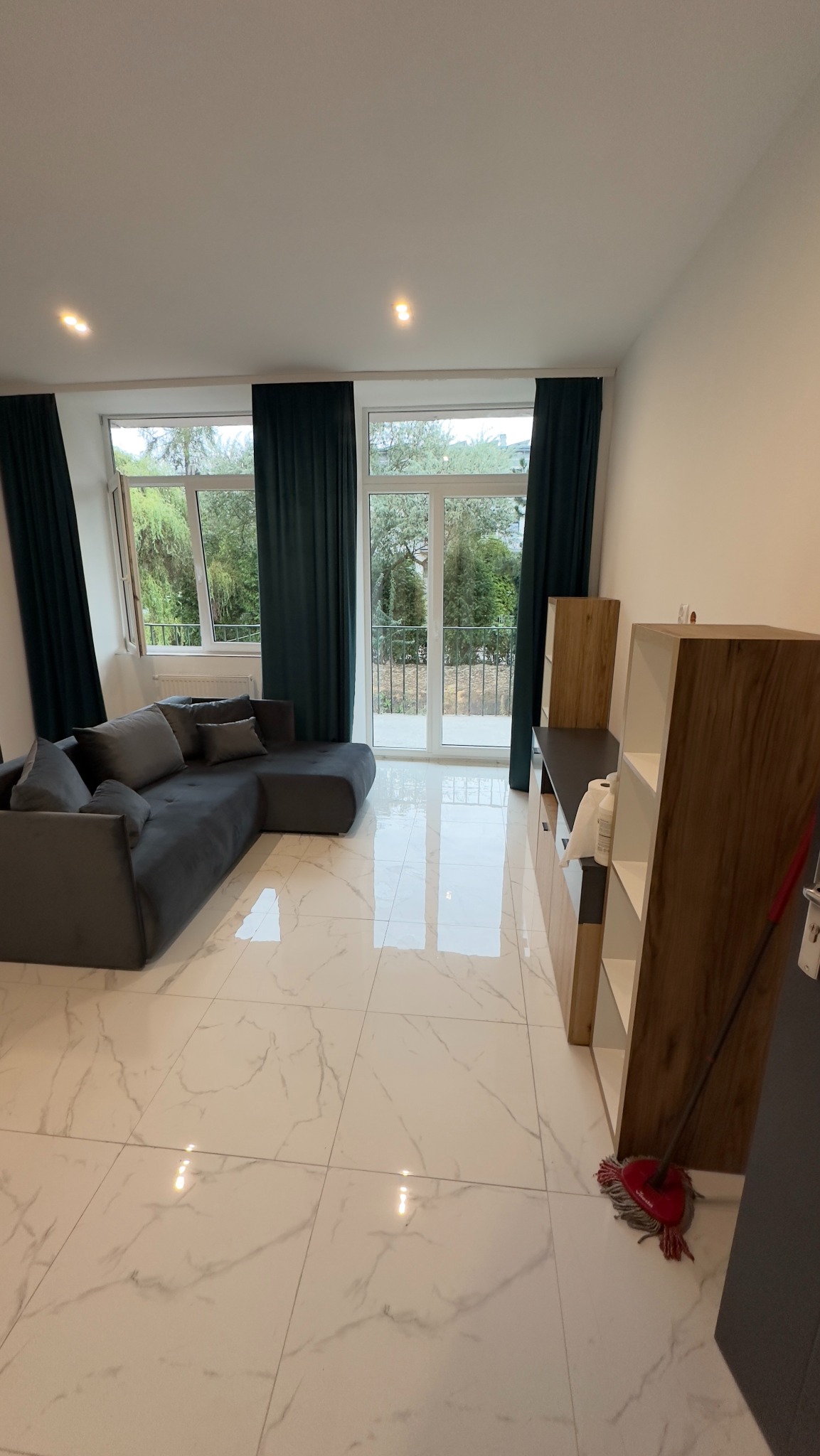Salon z szarym narożnikiem, marmurową podłogą i widokiem na balkon przez przeszklone drzwi balkonowe, z mopem stojącym w rogu pomieszczenia.