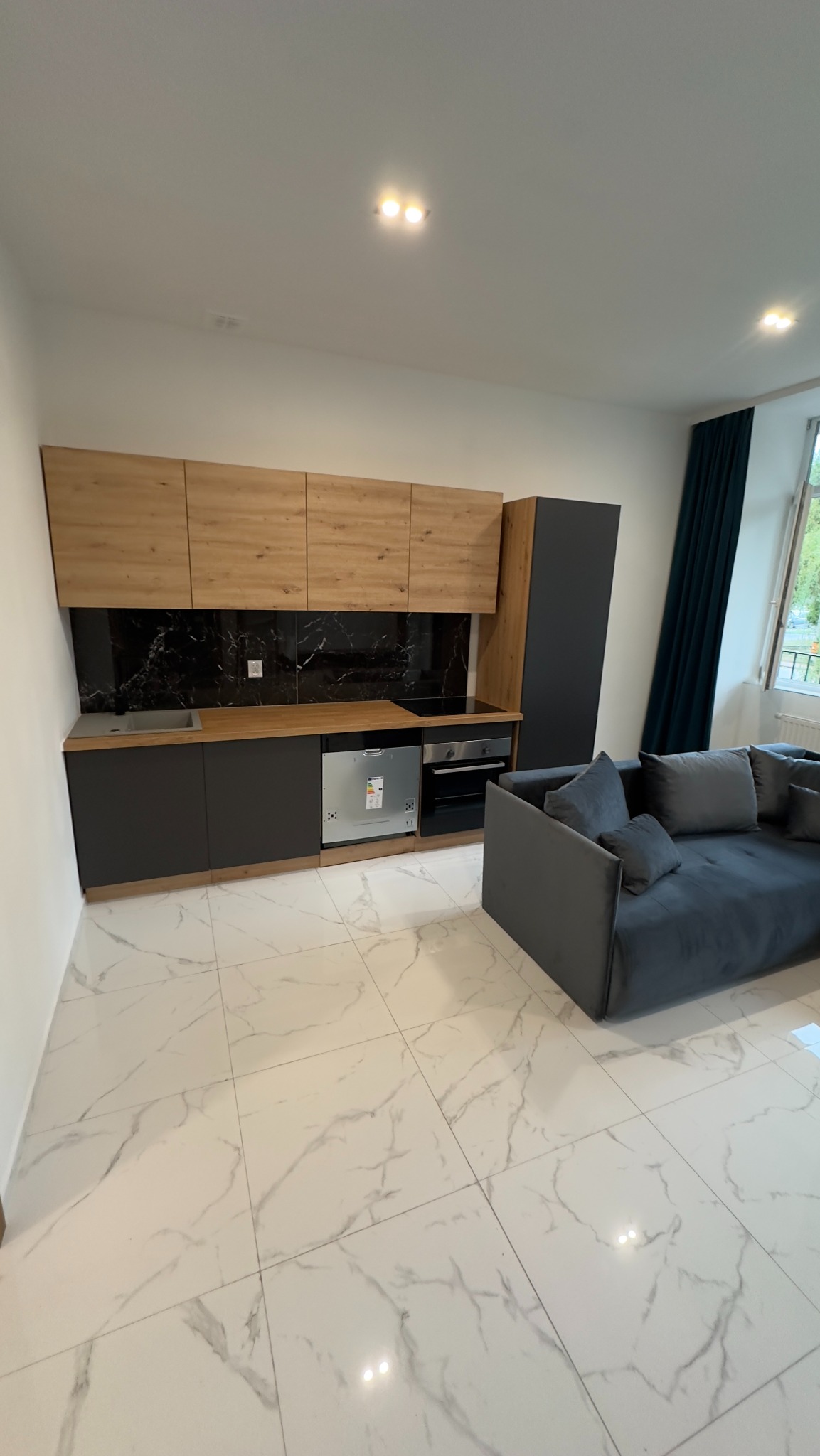 Nowoczesna kuchnia z drewnianymi szafkami górnymi i czarnymi szafkami dolnymi, marmurowym blatem i płytkami, zintegrowanym sprzętem AGD i szarą sofą w tle, na podłodze białe płytki imitujące marmur.