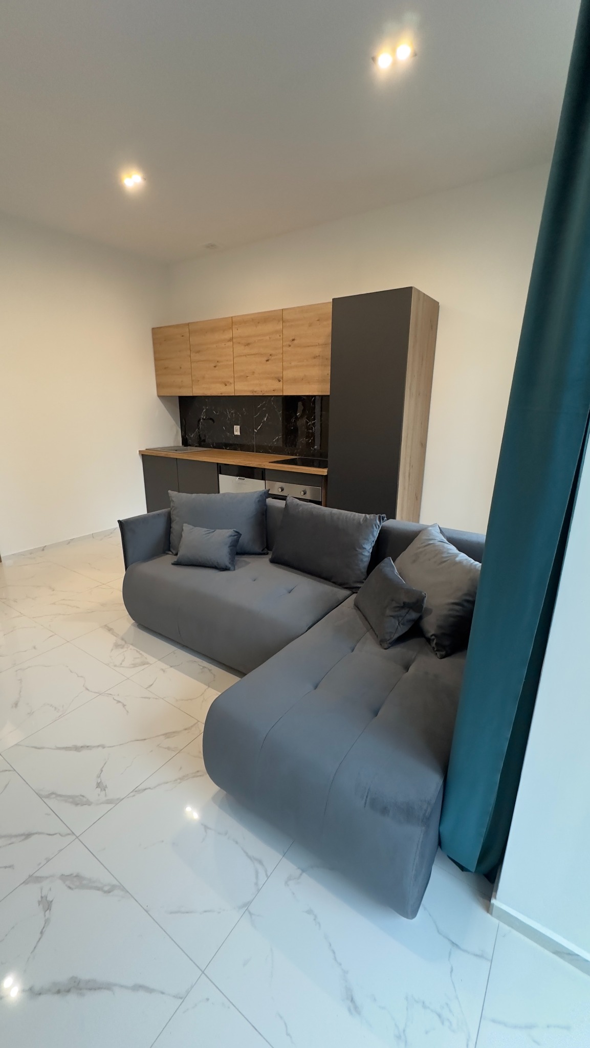 Wnętrze mieszkania z szarą sofą narożną i widoczną zabudową kuchenną w odcieniach drewna i grafitu, marmurowe płytki podłogowe.