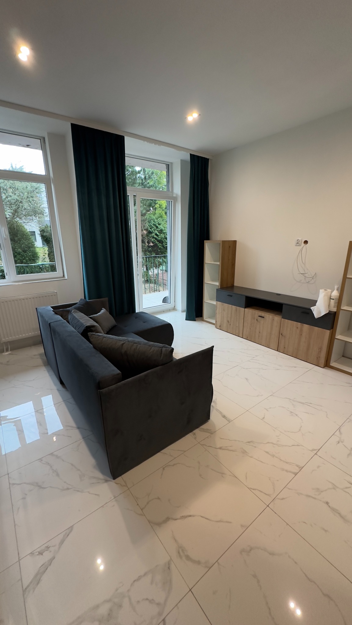 Nowoczesny salon z szarą sofą narożną, białymi marmurowymi płytkami podłogowymi, meblami RTV w odcieniach drewna i szarości oraz ciemnozielonymi zasłonami.