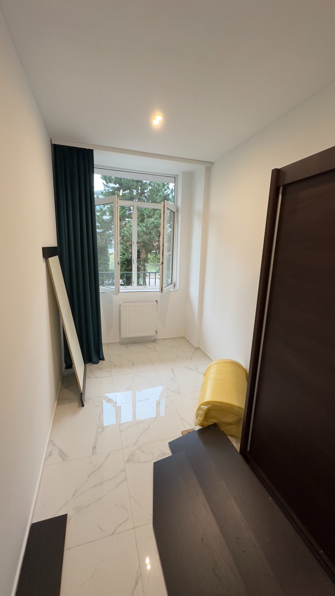 Pusty pokój z białymi ścianami, marmurową podłogą i otwartym oknem z widokiem na drzewa, przygotowany do prac wykończeniowych, z widocznymi materiałami budowlanymi i elementami drzwi.