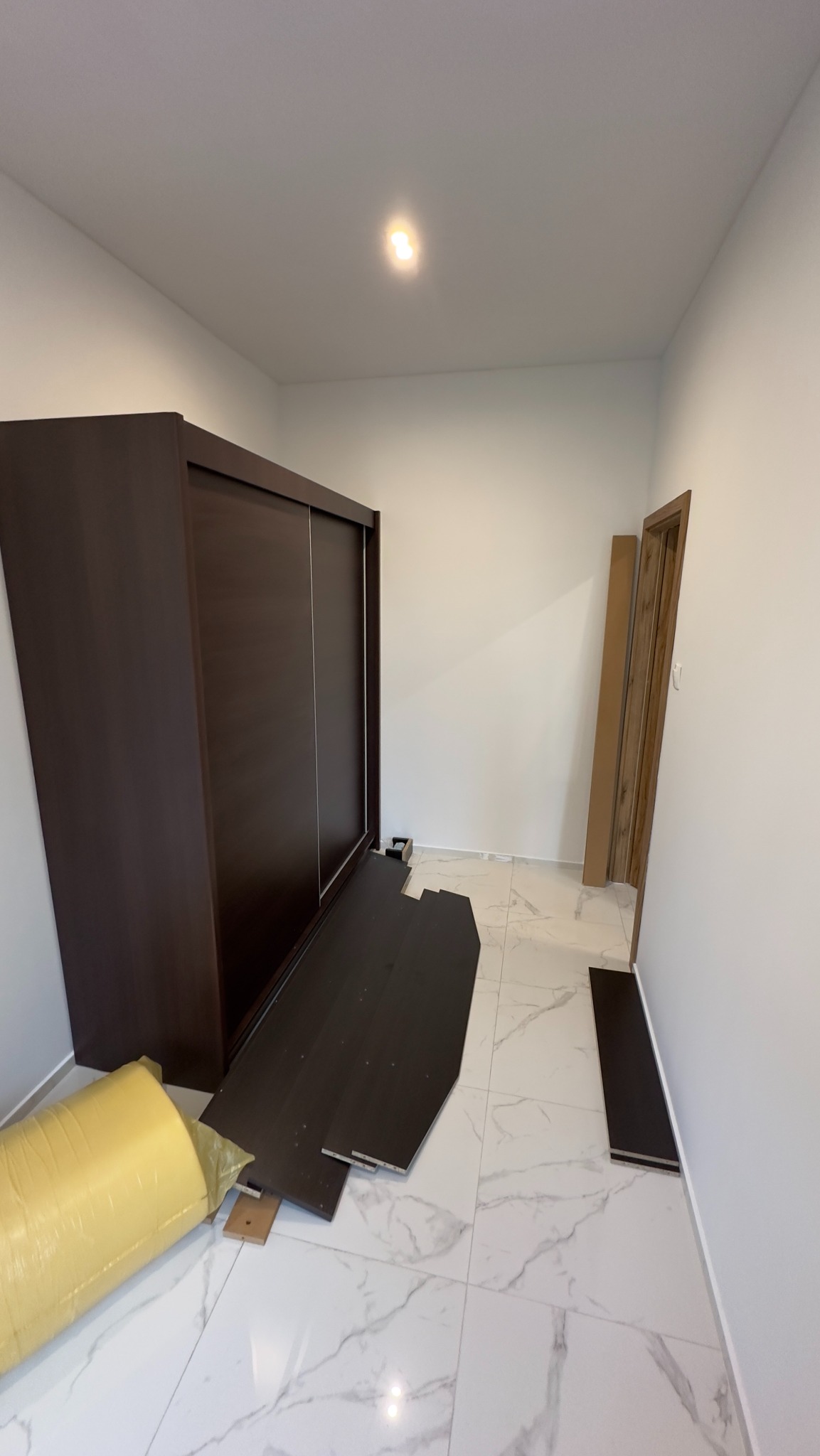 Rozpakowana, częściowo zmontowana, brązowa szafa przesuwna na marmurowej podłodze w pustym pokoju, widoczna pianka ochronna i elementy konstrukcyjne.