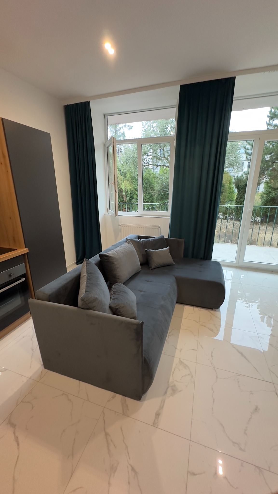 Szary, welurowy narożnik z poduszkami w salonie z białymi marmurowymi płytkami podłogowymi, ciemnozielonymi zasłonami i oknem z widokiem na zieleń.