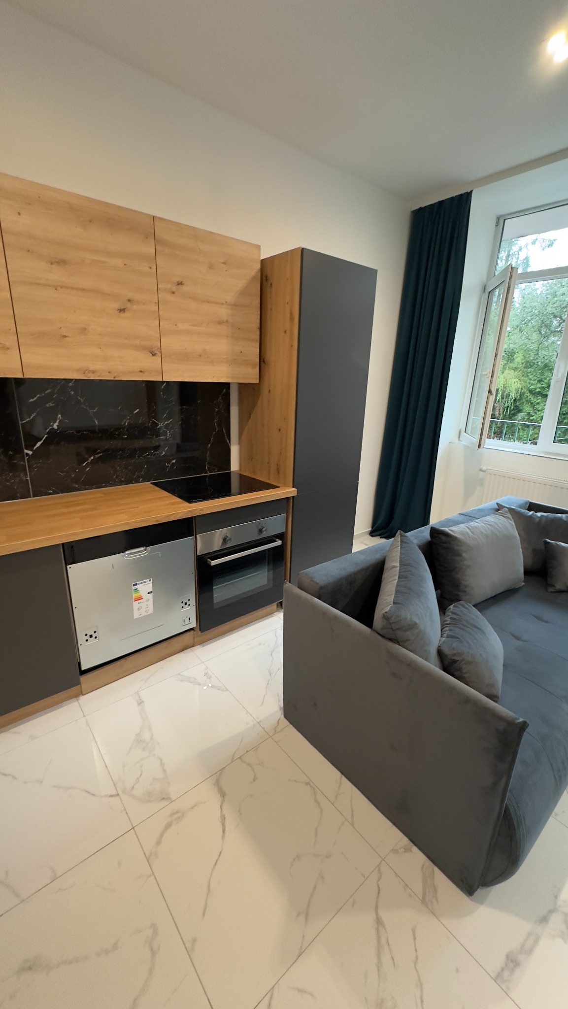 Nowoczesna kuchnia z drewnianymi frontami szafek, czarnym blatem i marmurową ścianą, widok na szarą zabudowę i narożnik z poduszkami, jasna marmurowa podłoga, okno z zielonymi zasłonami w tle.