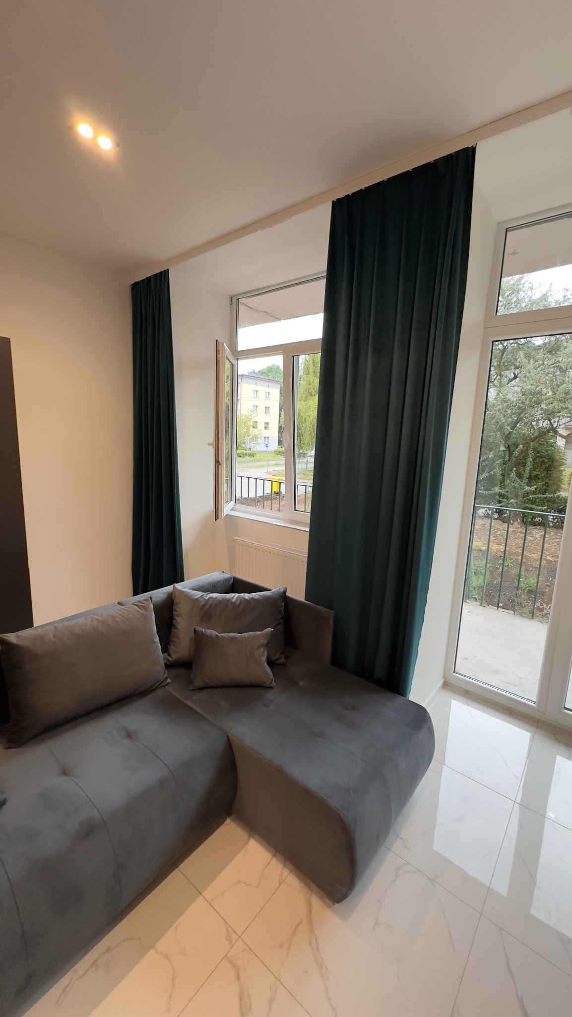 Szary narożnik z poduszkami w salonie z oknami przesłoniętymi długimi, ciemnozielonymi zasłonami, widok na zewnątrz przez uchylone okno.