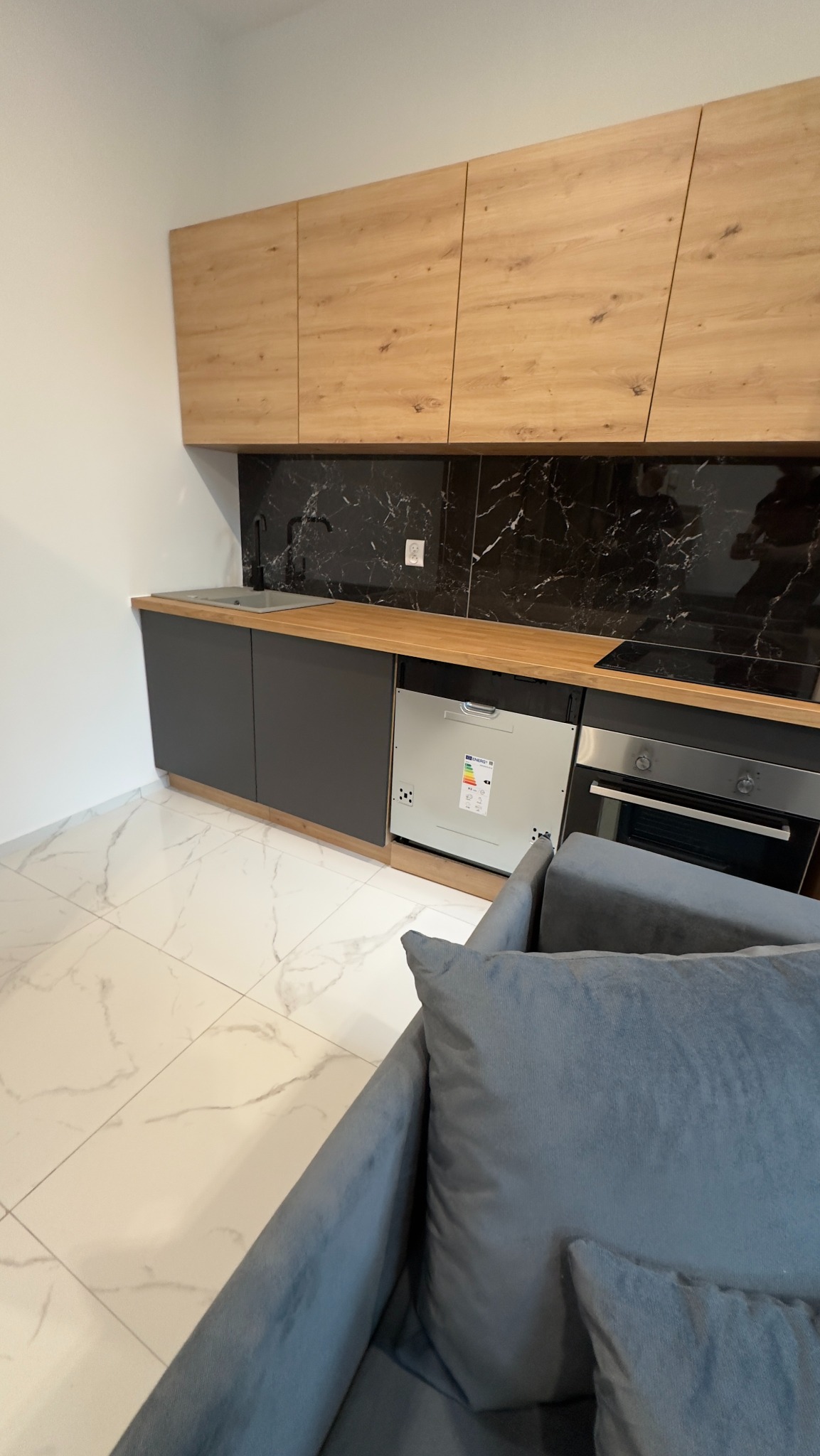 Nowoczesna kuchnia z drewnianymi szafkami górnymi, szarymi szafkami dolnymi i czarnym marmurowym backsplashiem, widok z perspektywy sofy.