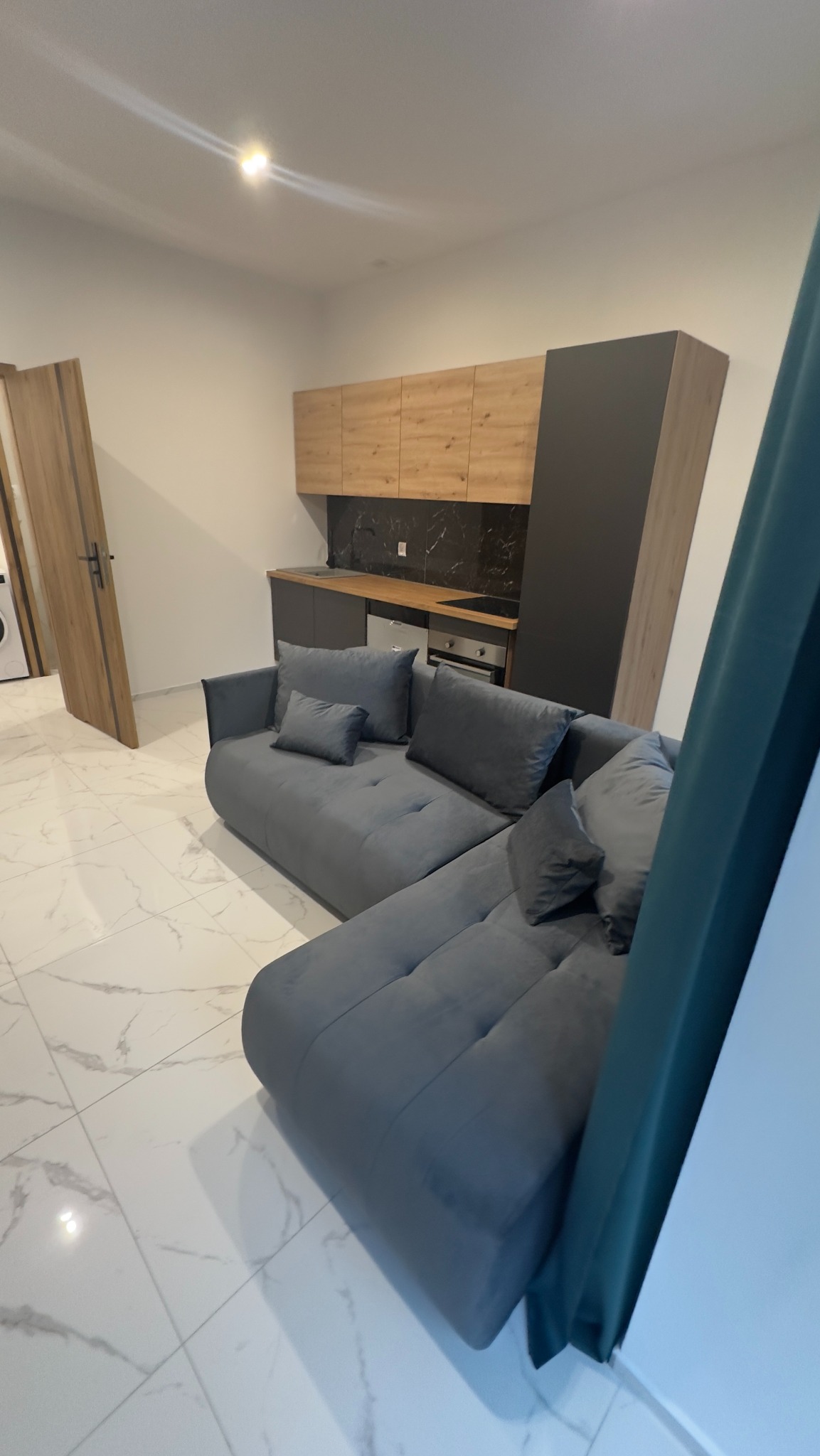 Nowoczesna kuchnia z szarymi szafkami i drewnianymi frontami w Bytomiu, widoczna z perspektywy narożnej sofy w odcieniach szarości na białej marmurowej podłodze.