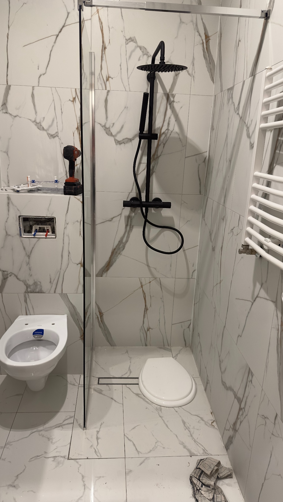 Nowoczesna łazienka w trakcie wykańczania: białe płytki imitujące marmur, czarna deszczownica, wisząca miska WC, grzejnik drabinkowy i narzędzia glazurnicze na ścianie.