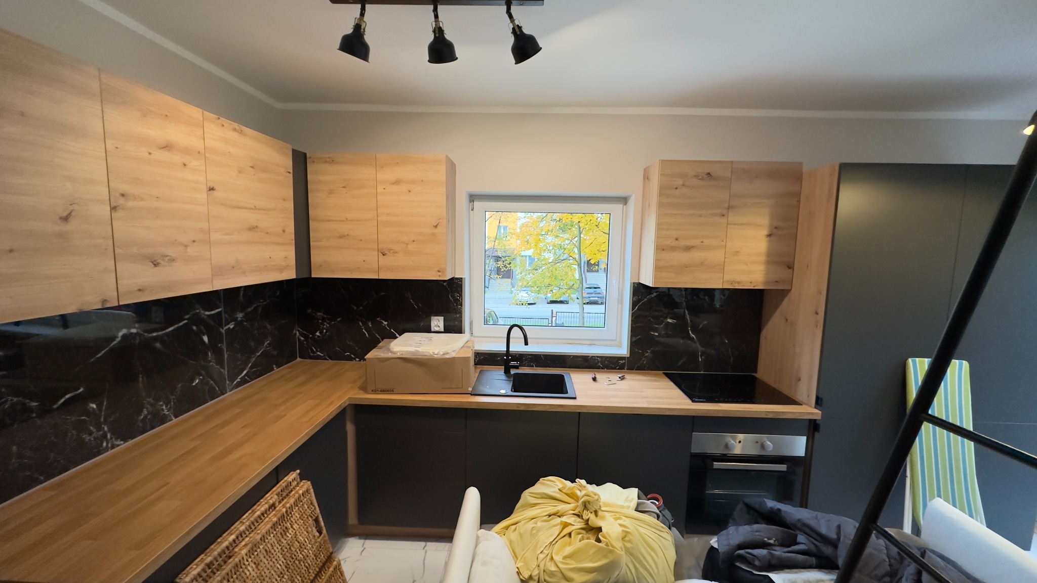 Nowoczesna kuchnia w trakcie montażu: drewniane szafki górne, czarny marmur na ścianie, czarne szafki dolne, okno z widokiem na jesienne drzewo, widoczne narzędzia i kartonowe opakowania.