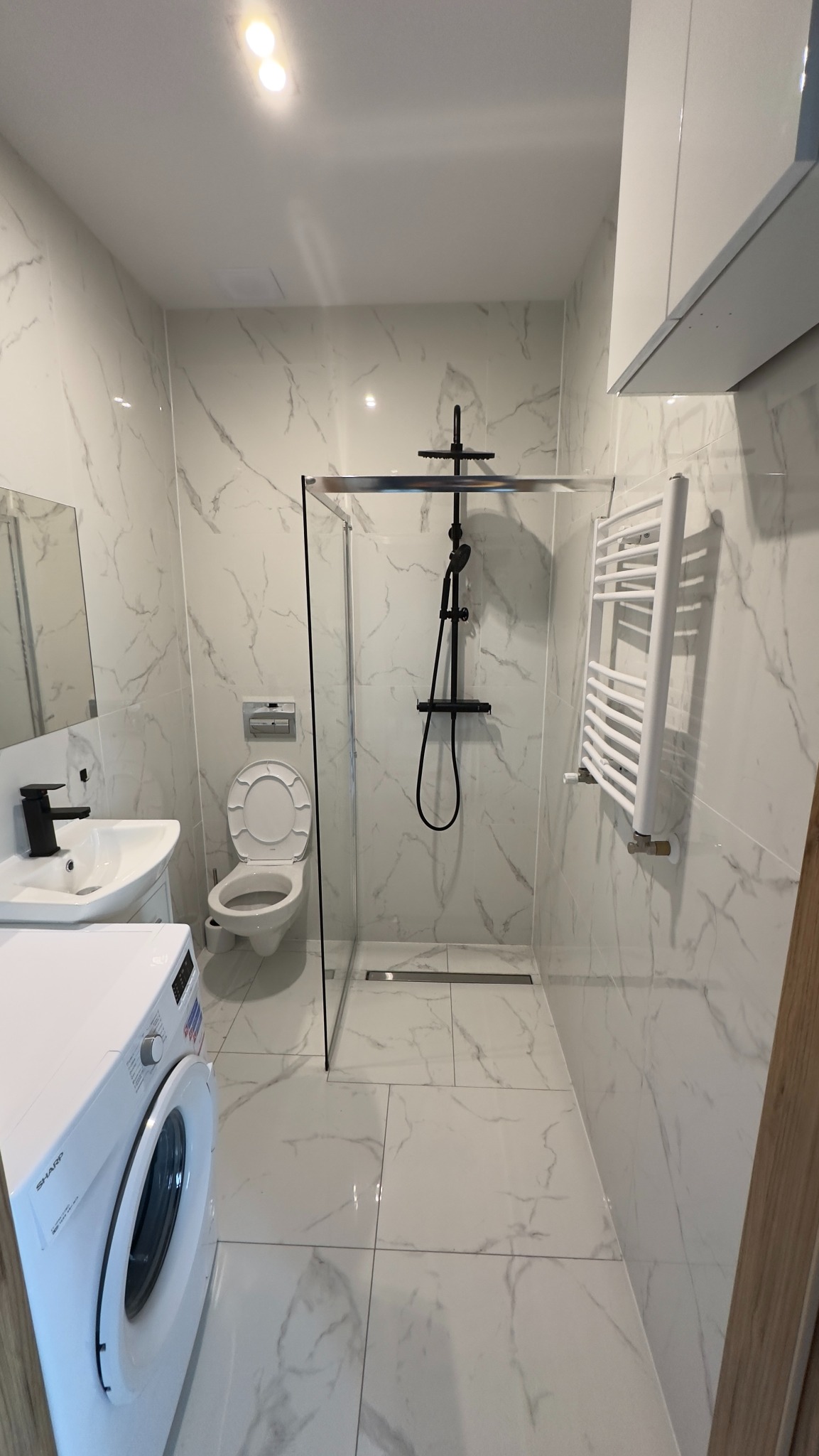 Biała łazienka z marmurowymi płytkami, kabiną prysznicową typu walk-in z czarną armaturą, toaletą, umywalką z czarną baterią i pralką Sharp.