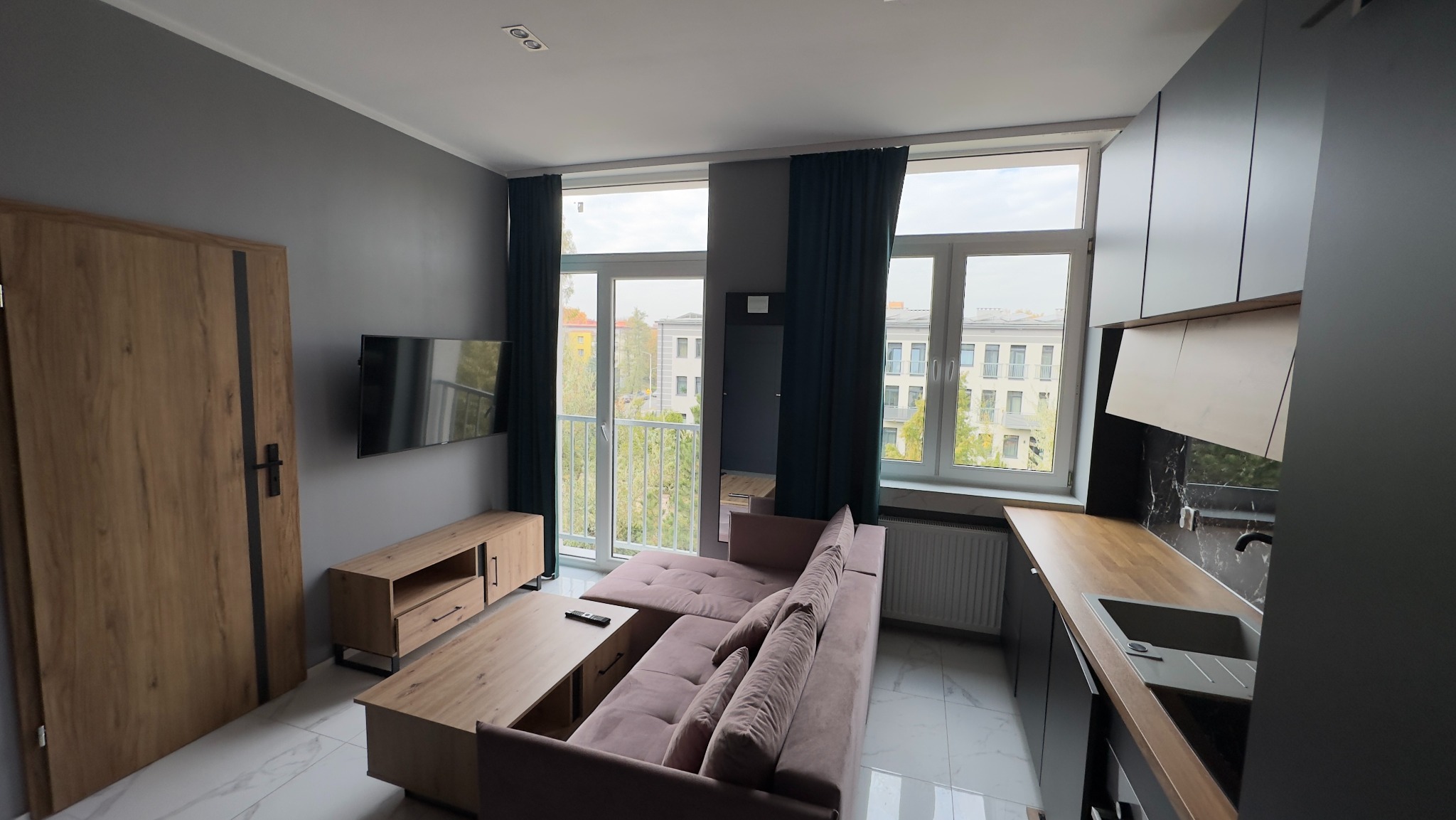 Nowoczesne wnętrze mieszkania z szarą ścianą, drewnianymi drzwiami z czarnym paskiem, telewizorem, szafką RTV, rozłożystą sofą w odcieniach różu, stolikiem kawowym, oknami z ciemnymi zasłonami...