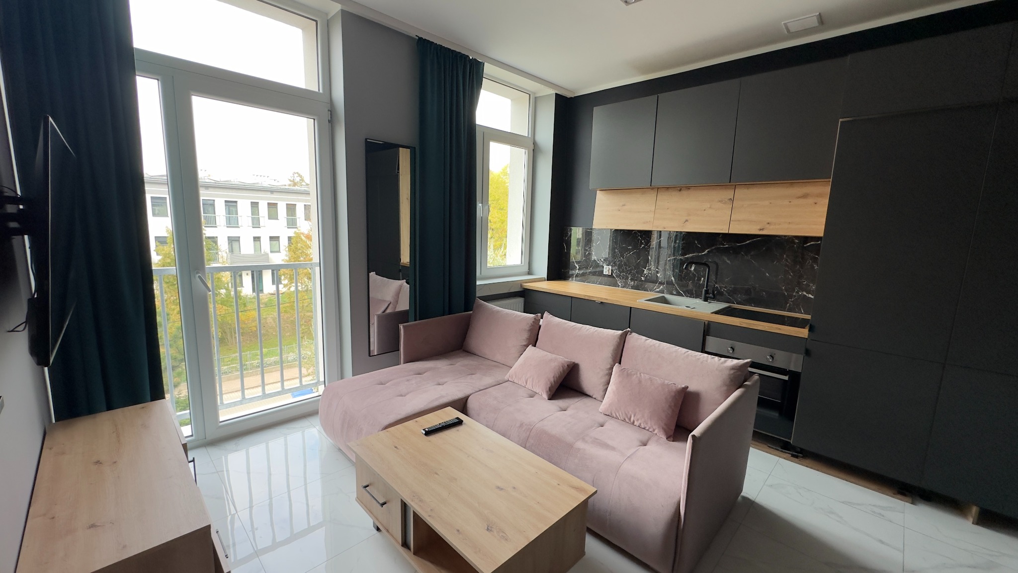 Nowoczesne wnętrze mieszkania z aneksem kuchennym: czarne szafki kuchenne z drewnianymi akcentami, marmurowy backsplash, jasnoróżowa sofa narożna, drewniany stolik kawowy i widok na balkon...