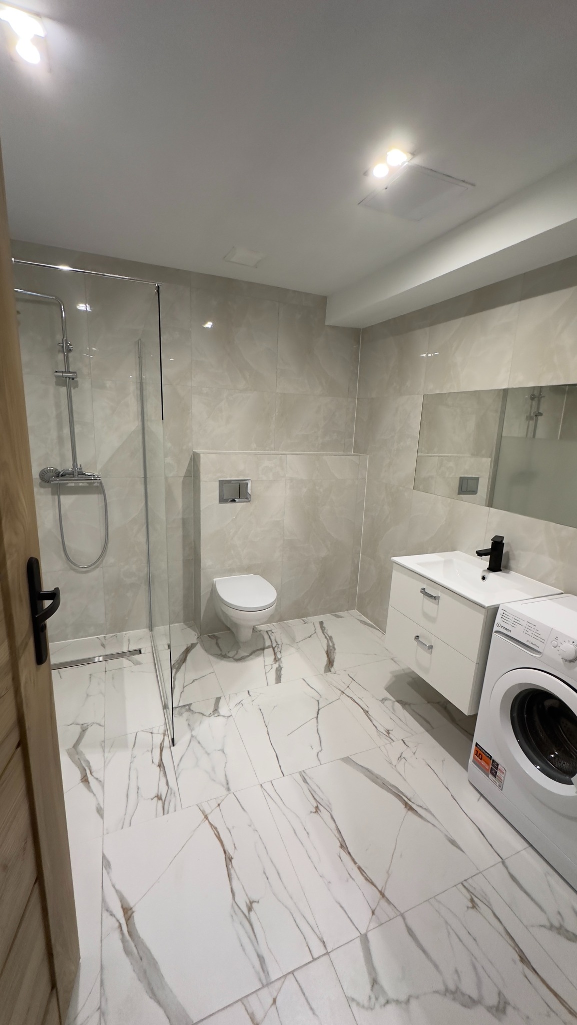 Jasna łazienka po remoncie z kabiną prysznicową typu walk-in, toaletą podwieszaną, białą szafką z umywalką i pralką, wykończona płytkami imitującymi marmur.