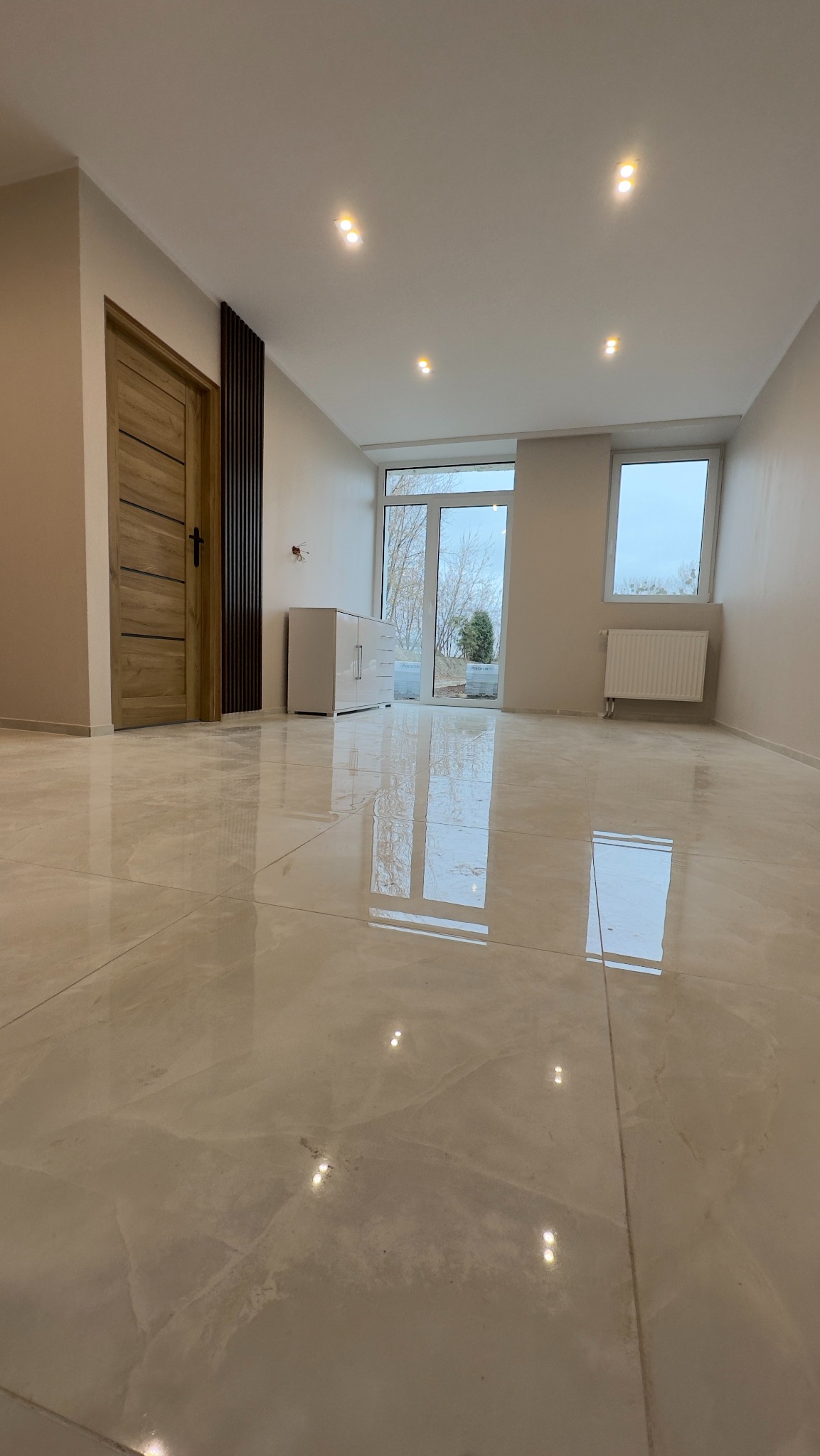 Jasne, przestronne wnętrze z połyskującą podłogą z płytek imitujących marmur, drewnianymi drzwiami z poziomymi panelami i białymi meblami, doświetlone punktowymi lampami sufitowymi.