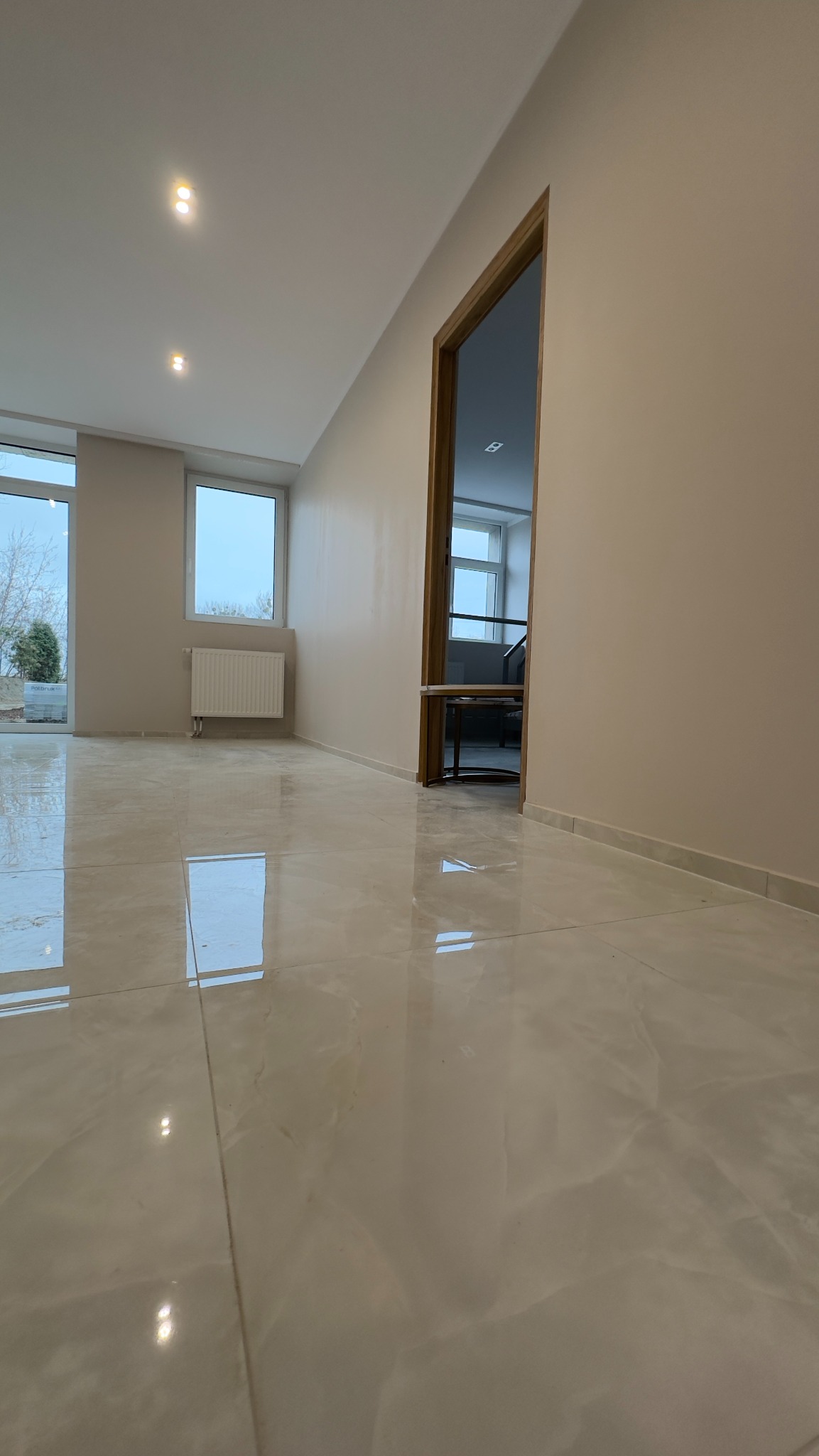 Jasne, błyszczące płytki podłogowe imitujące marmur w nowym, pustym pokoju z beżowymi ścianami, widoczne okna i drzwi do kolejnego pomieszczenia.