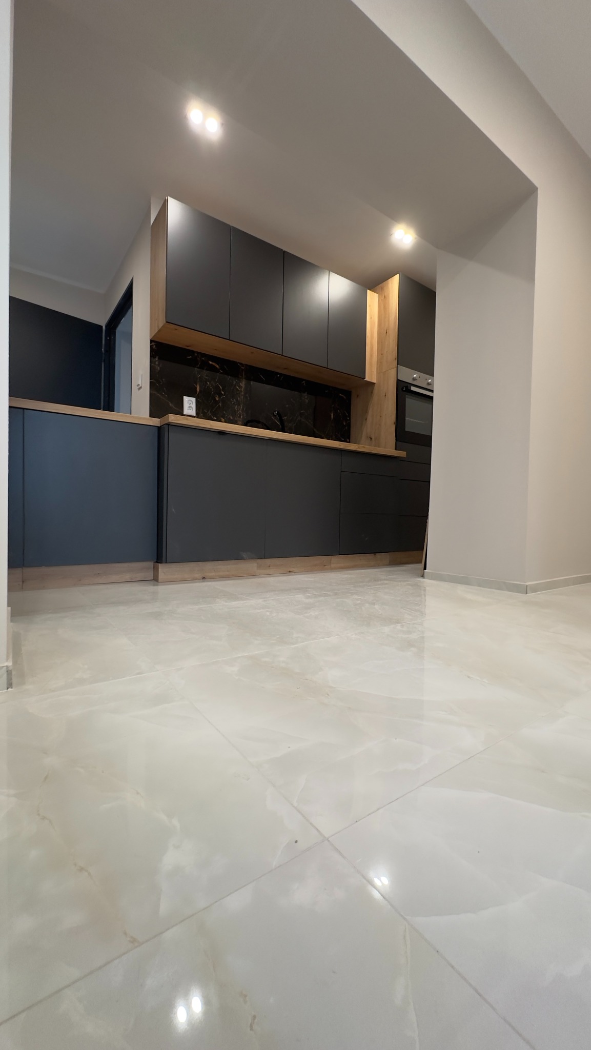 Nowoczesna kuchnia z szarymi szafkami, drewnianymi akcentami i marmurowym backsplashiem, oświetlona punktowymi lampami, z jasną marmurową podłogą.