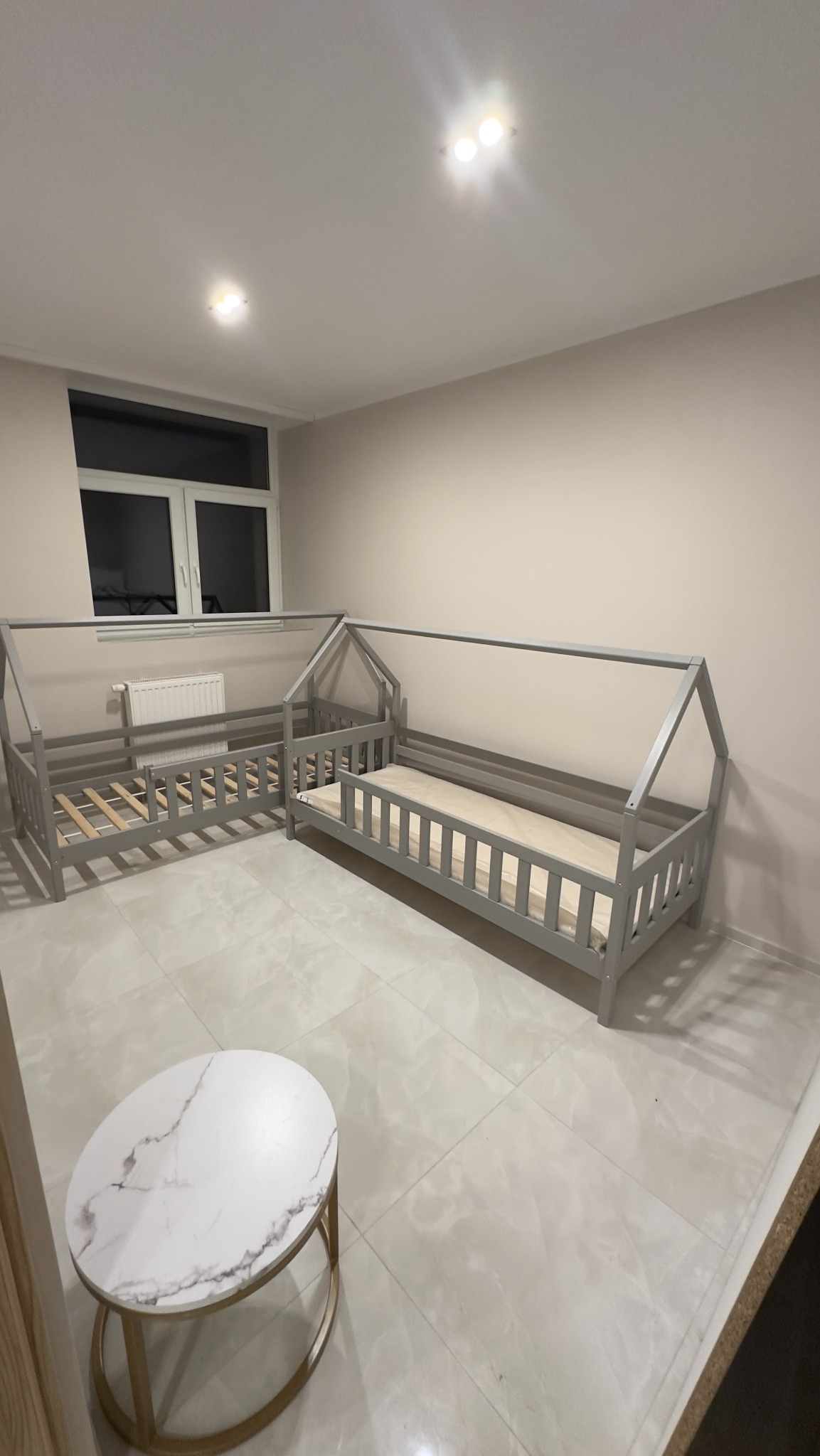 Dwa szare, drewniane łóżka dziecięce w kształcie domków, jedno z materacem, drugie z widocznym stelażem, ustawione na błyszczącej, jasnej podłodze w pokoju z oknem i małym stolikiem.