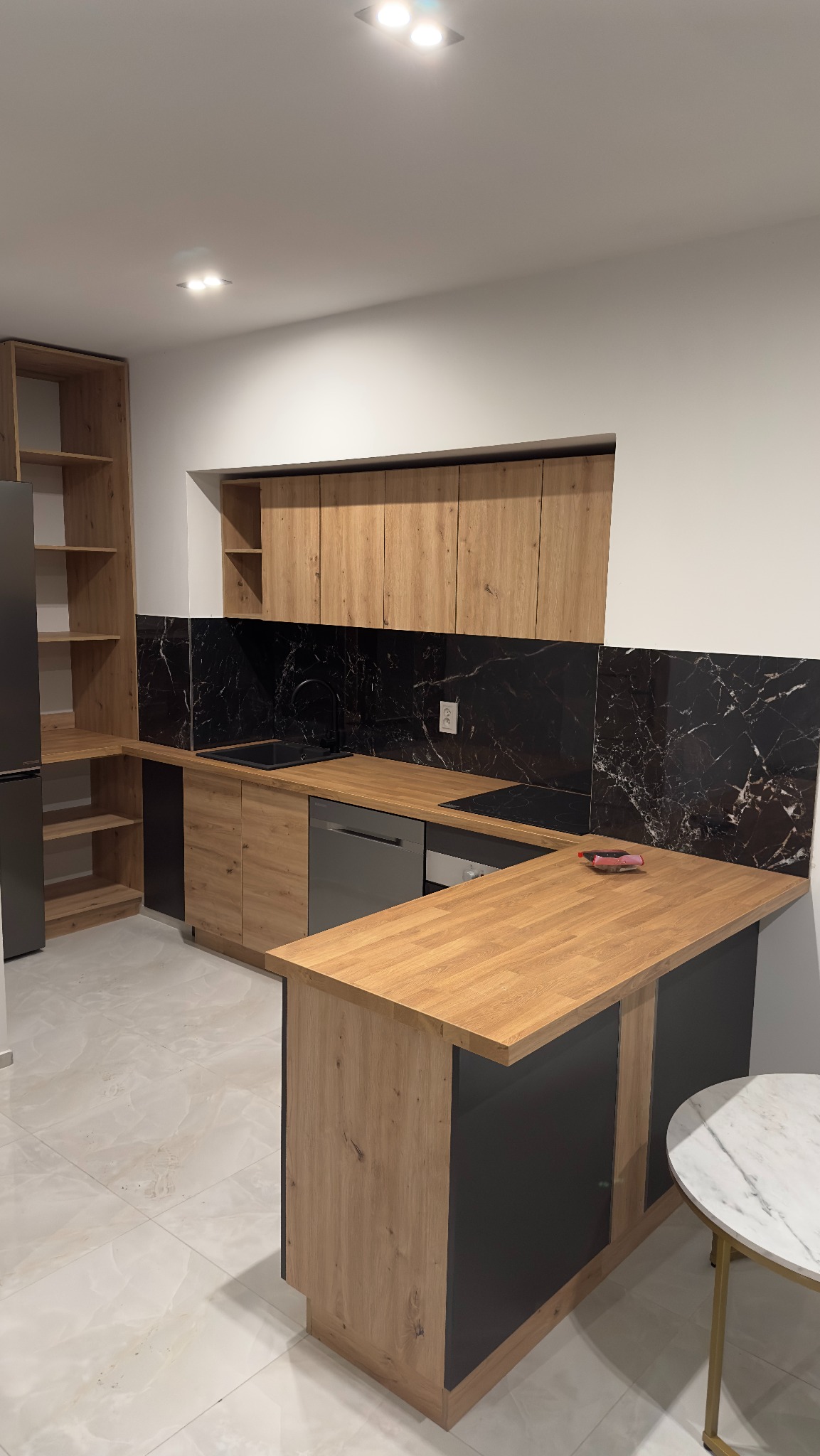 Nowoczesna kuchnia z drewnianymi szafkami i blatem, czarnym marmurowym backsplashiem oraz wyspą kuchenną z czarnymi frontami, widok na okrągły stolik z marmurowym blatem.