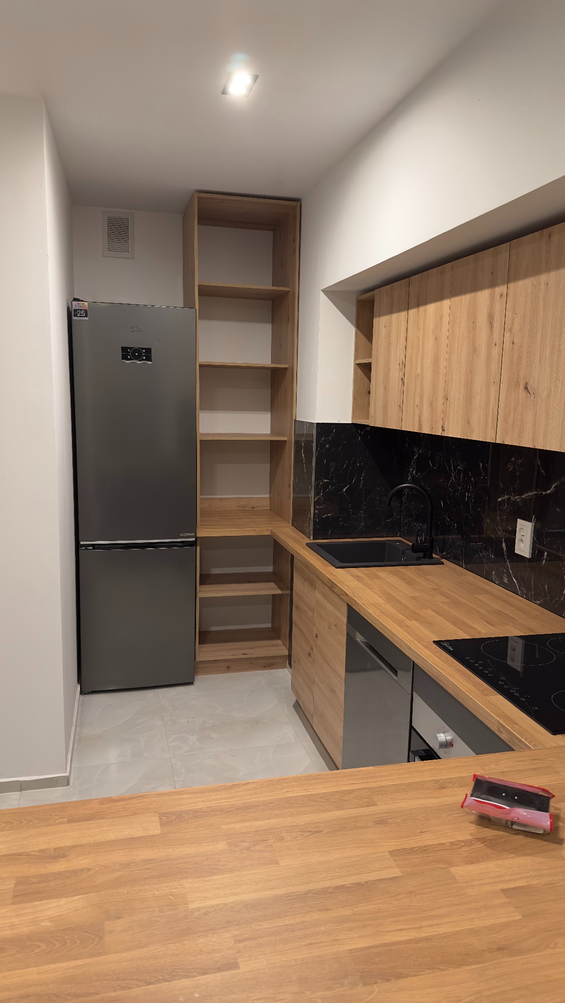 Nowoczesna kuchnia z drewnianymi szafkami, czarnym blatem i marmurowym backsplashiem, srebrna lodówka i drewniany barek z przodu.