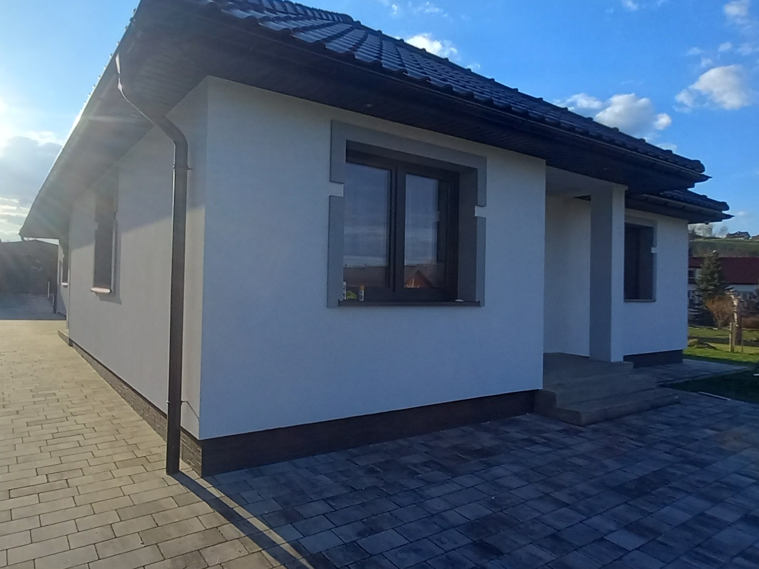 Nowoczesny dom jednorodzinny w Chełmcu z jasną elewacją, ciemną dachówką i szarą kostką brukową, ukazujący staranne wykonanie detali.