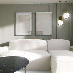 The Plan sp. z o.o. - Minimalistyczny salon z białym narożnikiem, okrągłym stolikiem z czarnego marmuru i abstrakcyjnymi obrazami w białych ramach na oliwkowozielonej ścianie.