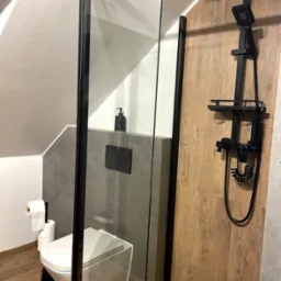 Nowoczesna kabina prysznicowa z czarnymi detalami i drewnopodobnymi płytkami w Zabrzedu. Umywalka podwieszana i minimalistyczna armatura.