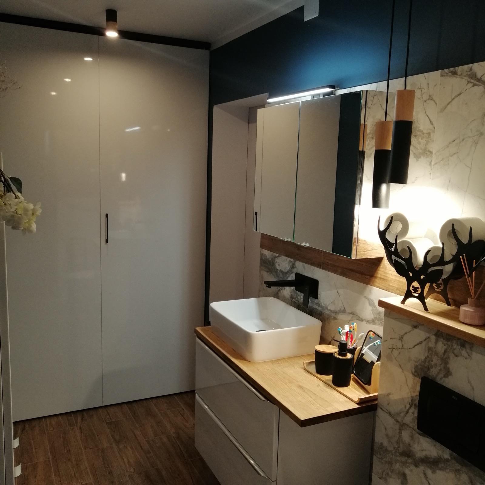 Nowoczesna łazienka z białą, prostokątną umywalką nablatową, czarną armaturą, szafką pod umywalką z drewnianym blatem, marmurową ścianą i dekoracyjnym czarnym reniferem na półce.