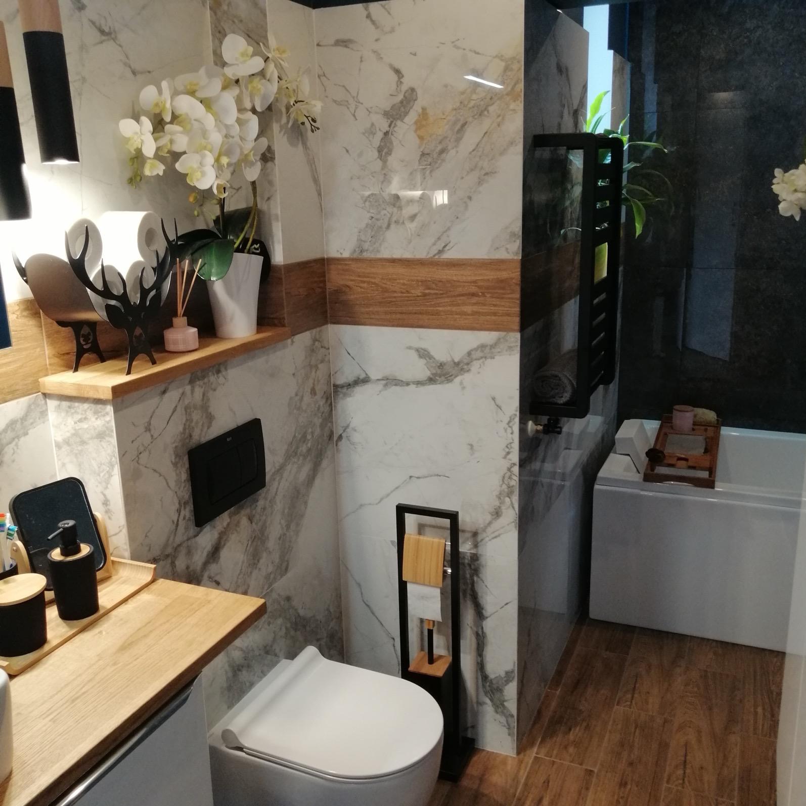 Nowoczesna łazienka z marmurowymi płytkami, drewnianymi akcentami, białą armaturą i dekoracyjnymi elementami, takimi jak sztuczne orchidee i czarny stojak na papier toaletowy.