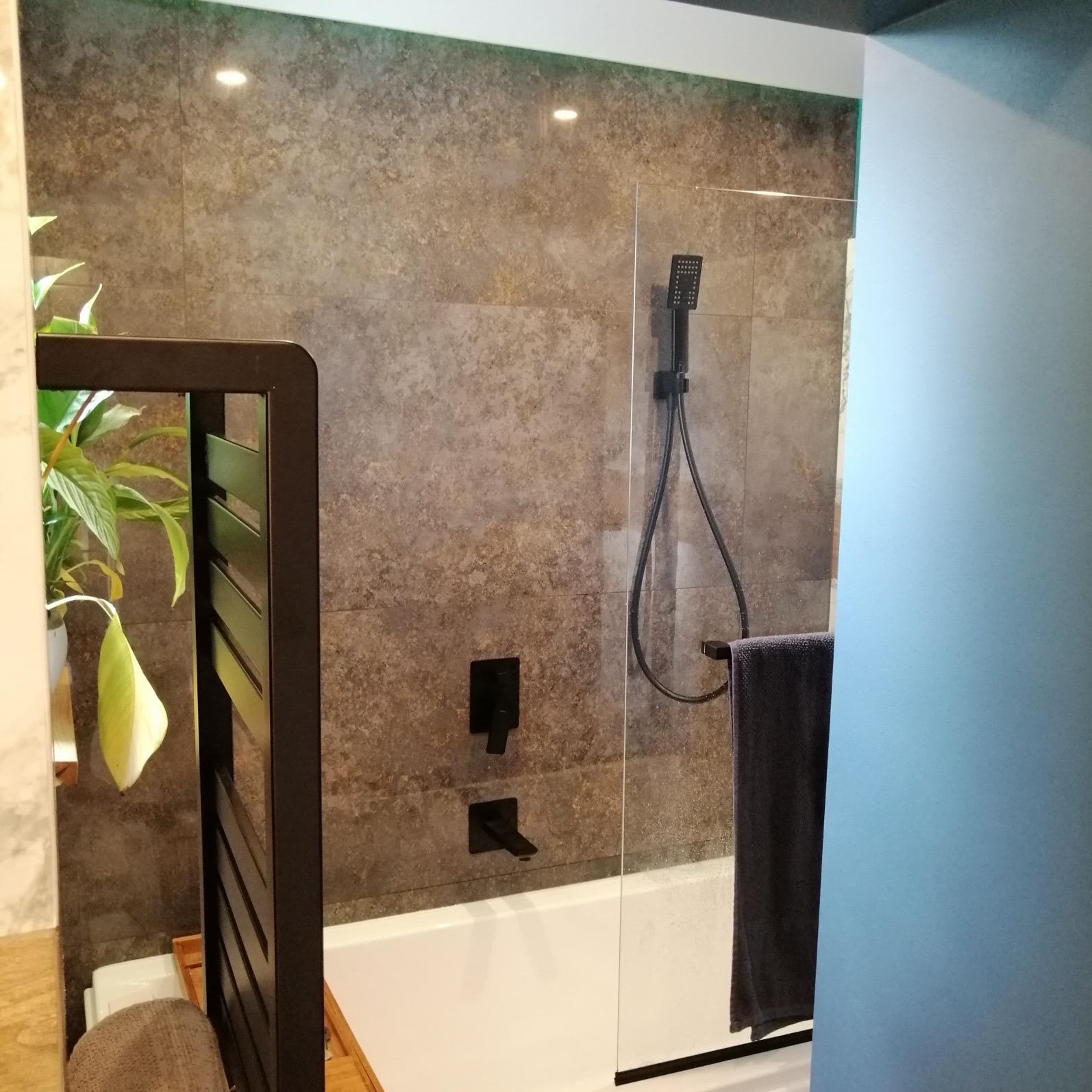 Nowoczesna łazienka z wanną, przeszkloną kabiną prysznicową, ciemnymi płytkami imitującymi kamień, czarną armaturą i dekoracyjnym parawanem z rośliną doniczkową.