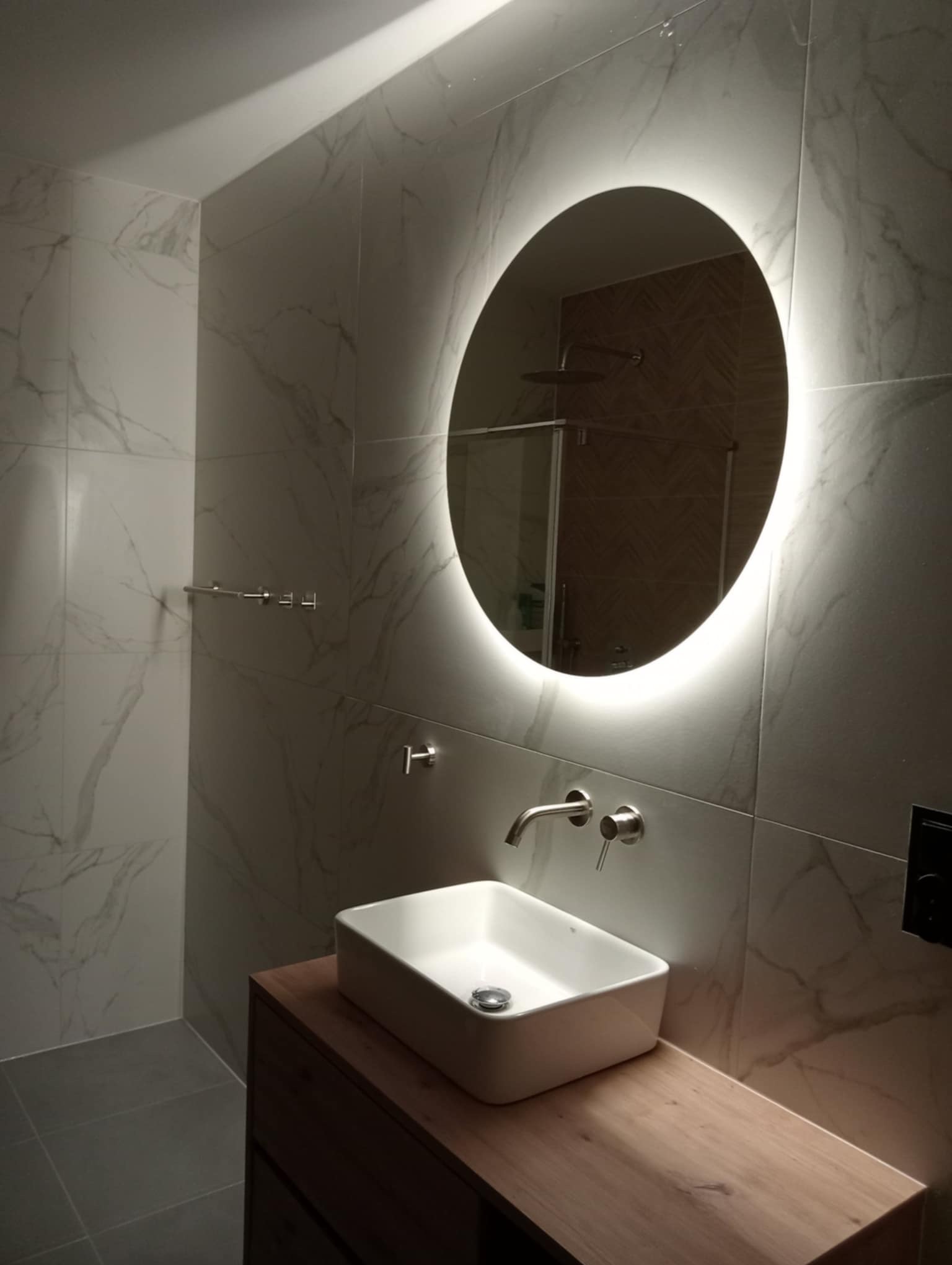Nowoczesna łazienka z owalnym lustrem LED, umywalką nablatową i marmurowymi płytkami. Widoczna armatura i fragment kabiny prysznicowej.