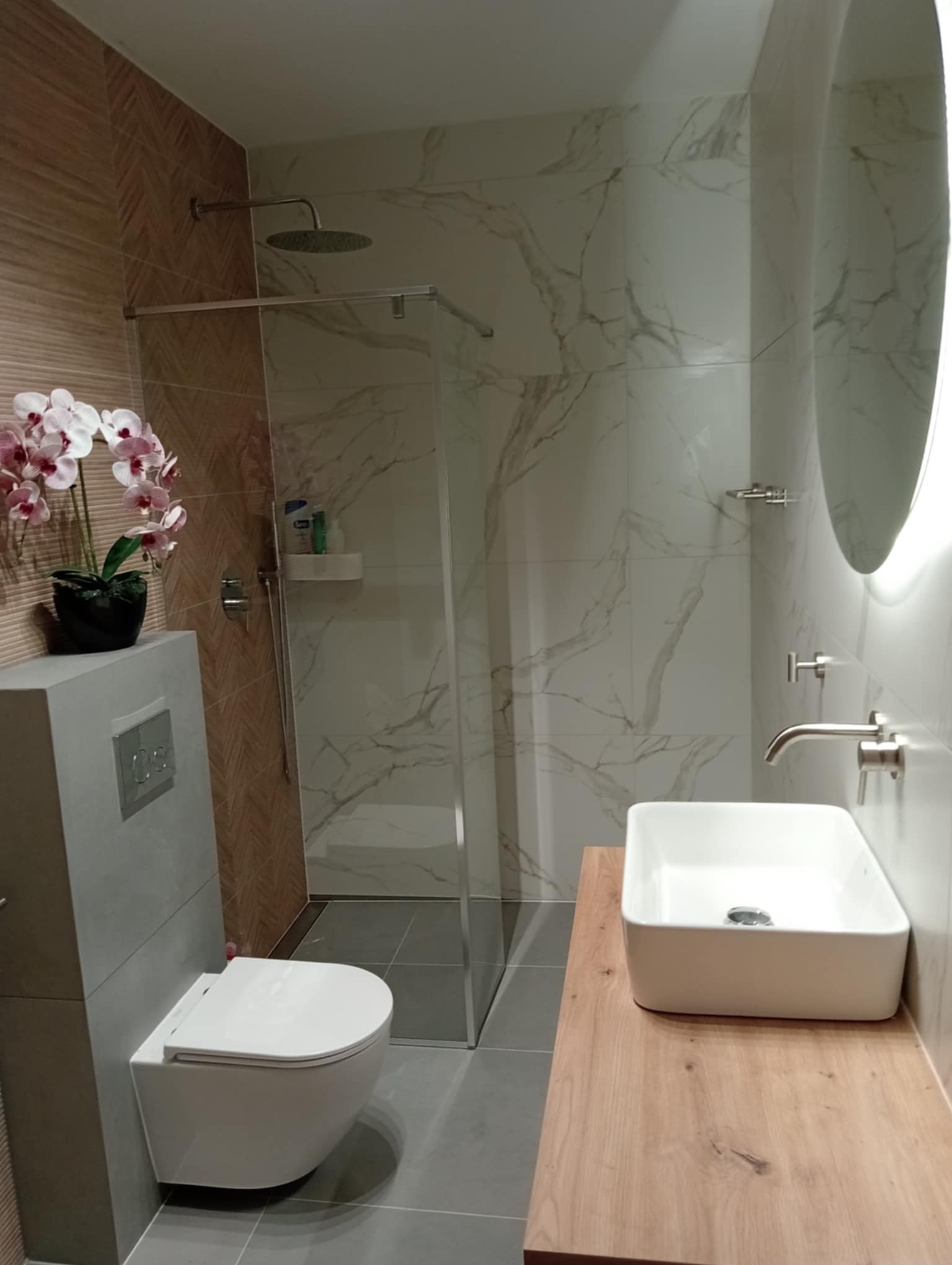 Nowoczesna łazienka z kabiną prysznicową typu walk-in, umywalką na drewnianym blacie i podwieszaną toaletą. Na ścianach płytki imitujące marmur i drewno.