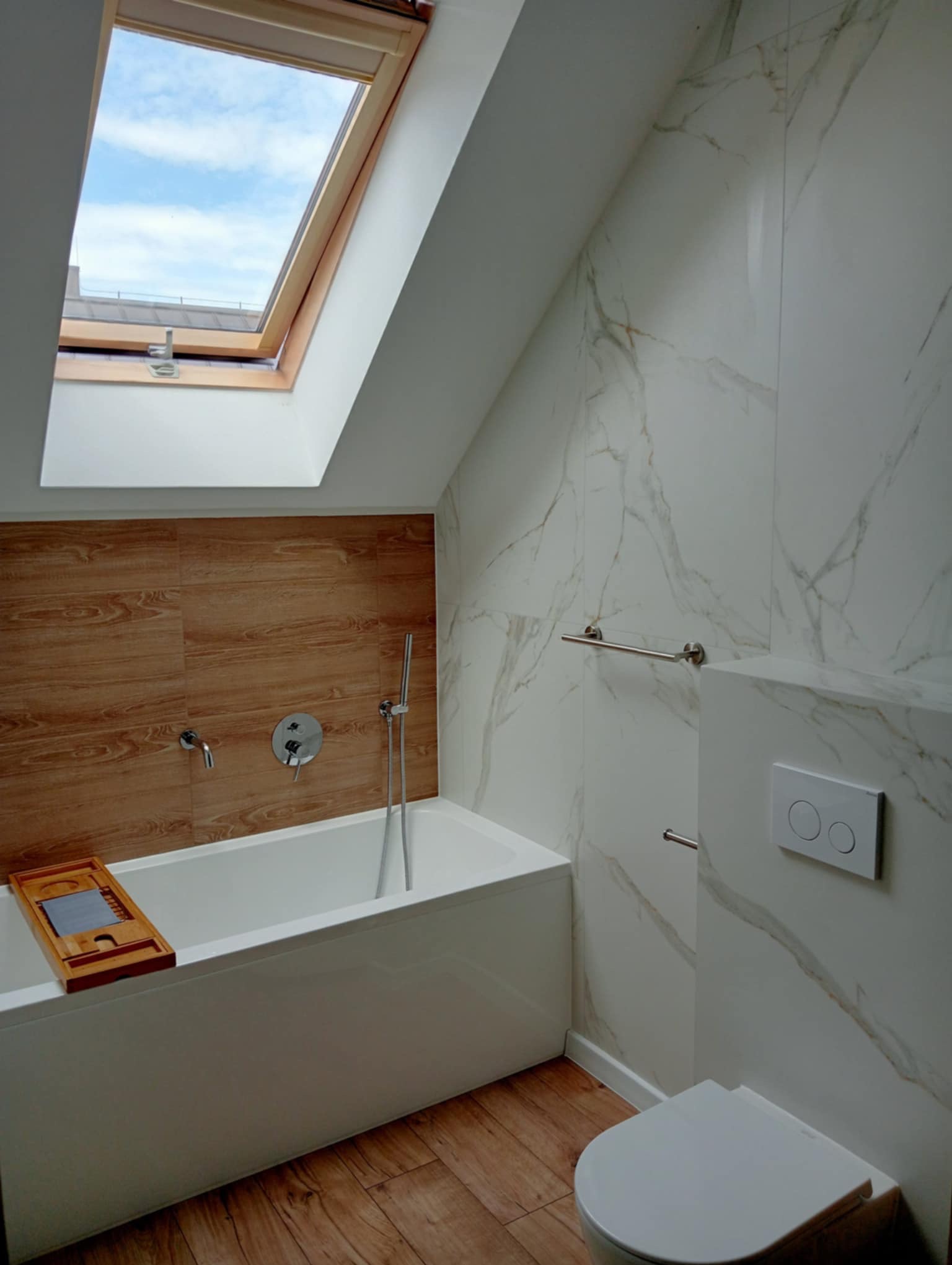 Nowoczesna łazienka na poddaszu z oknem dachowym, białą wanną i toaletą. Ściany wyłożone płytkami imitującymi marmur i drewno.