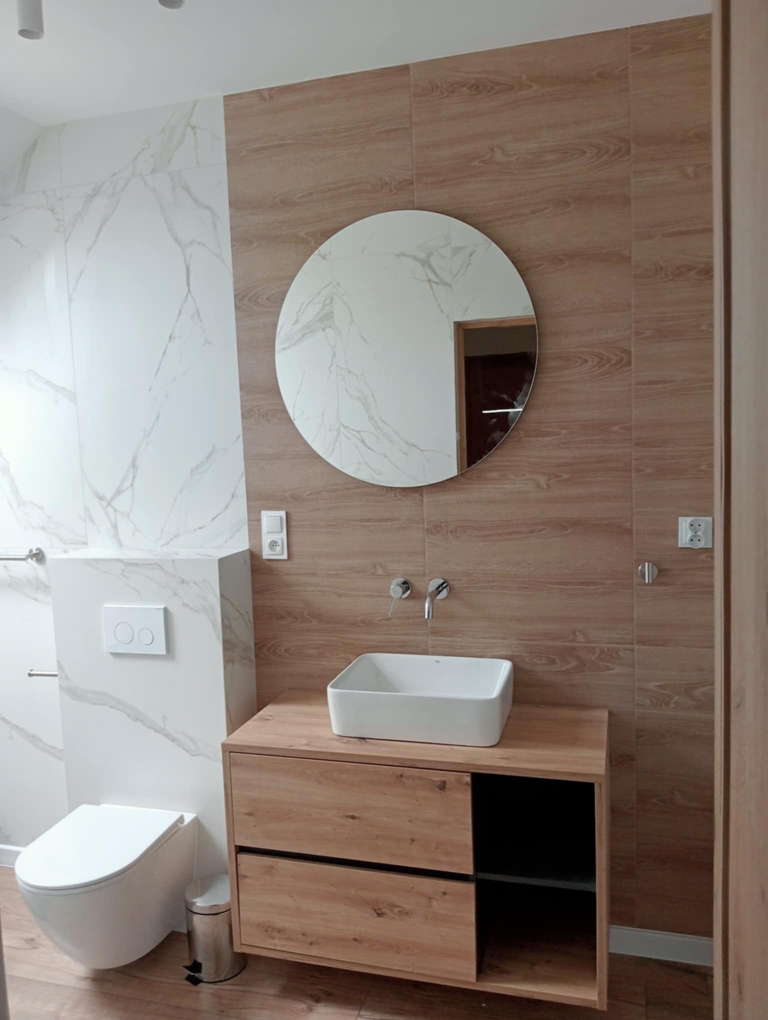 Nowoczesna łazienka z okrągłym lustrem i drewnopodobnymi płytkami ściennymi. Umywalka nablatowa na szafce z drewnianym frontem. Biała armatura i marmurowe płytki.