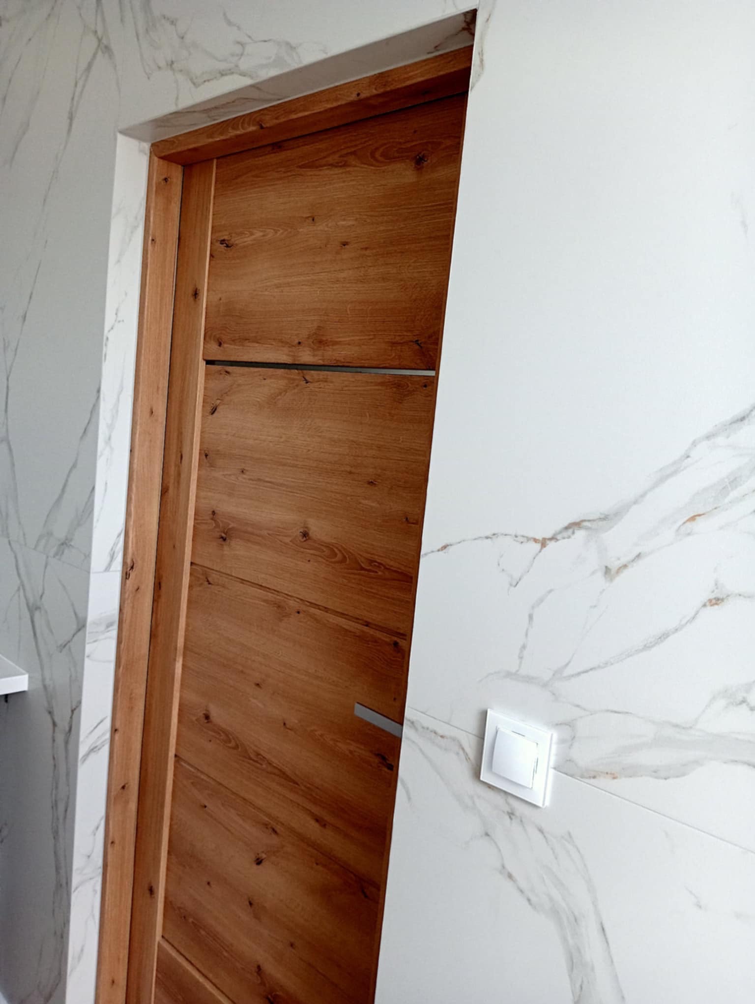 Wykończona łazienka: drewniane drzwi z poziomymi panelami i metalowym uchwytem, marmurowe płytki na ścianach, biały włącznik światła.