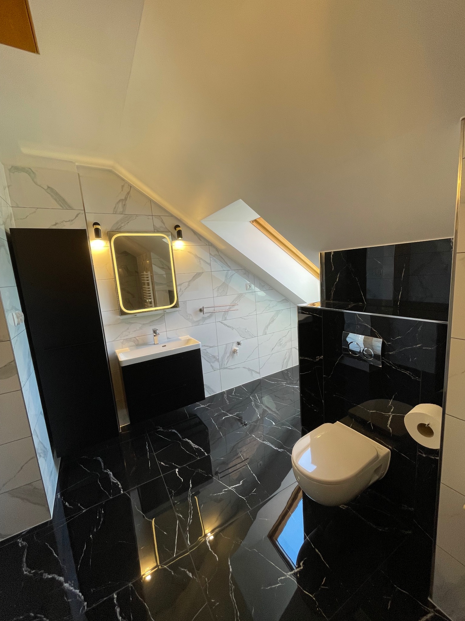 Nowoczesna łazienka na poddaszu z czarno-białymi płytkami imitującymi marmur, podwieszana toaleta i lustro z podświetleniem LED. Skosy i okno dachowe.