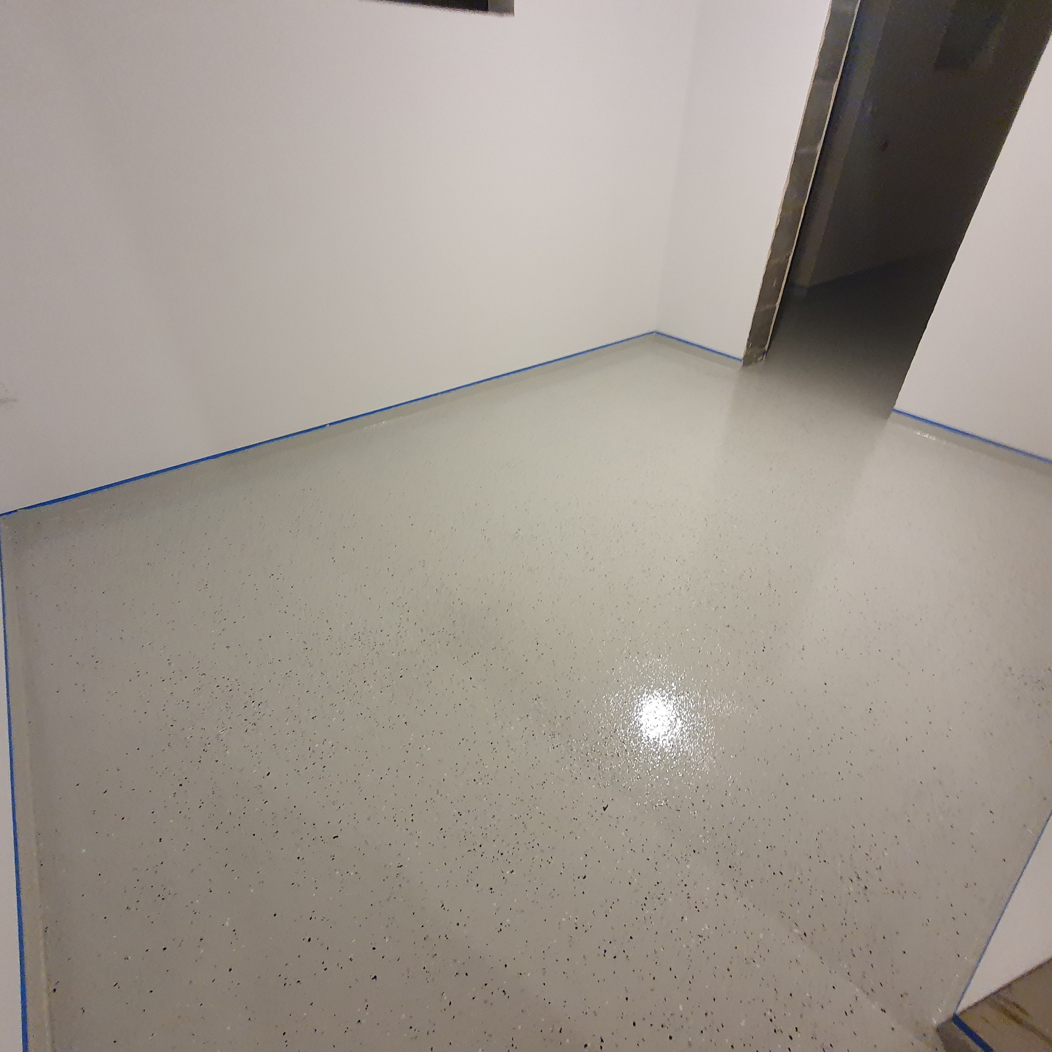 Świeżo wylana, jasnoszara posadzka żywiczna z drobnymi, ciemnymi inkluzjami w rogu pomieszczenia, zabezpieczona niebieską taśmą malarską przy ścianach.