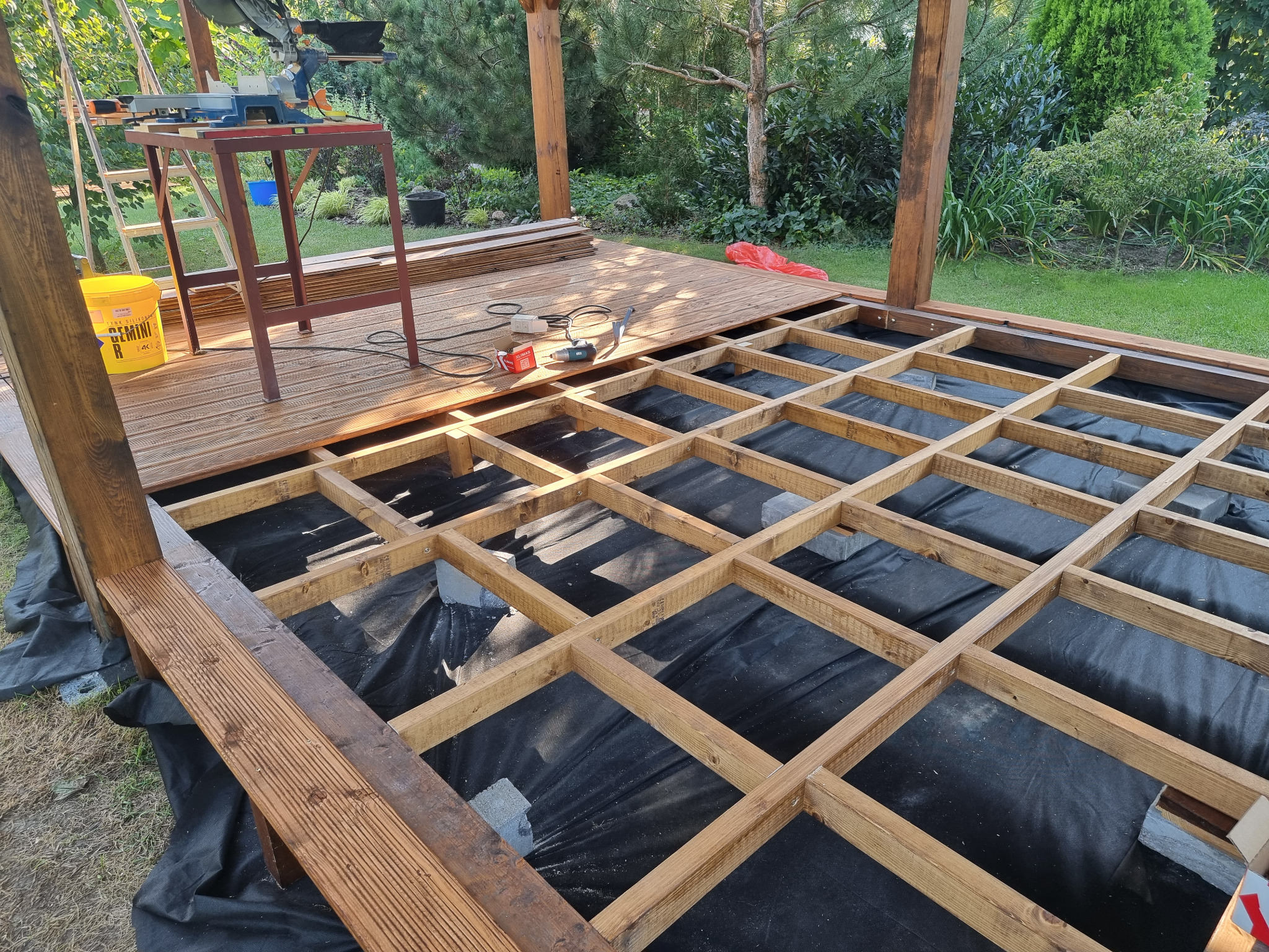 Konstrukcja drewnianego tarasu w ogrodzie na etapie budowy: widoczna podkonstrukcja z belek na czarnej geowłókninie, częściowo ułożone deski, narzędzia i piła kątowa w tle.