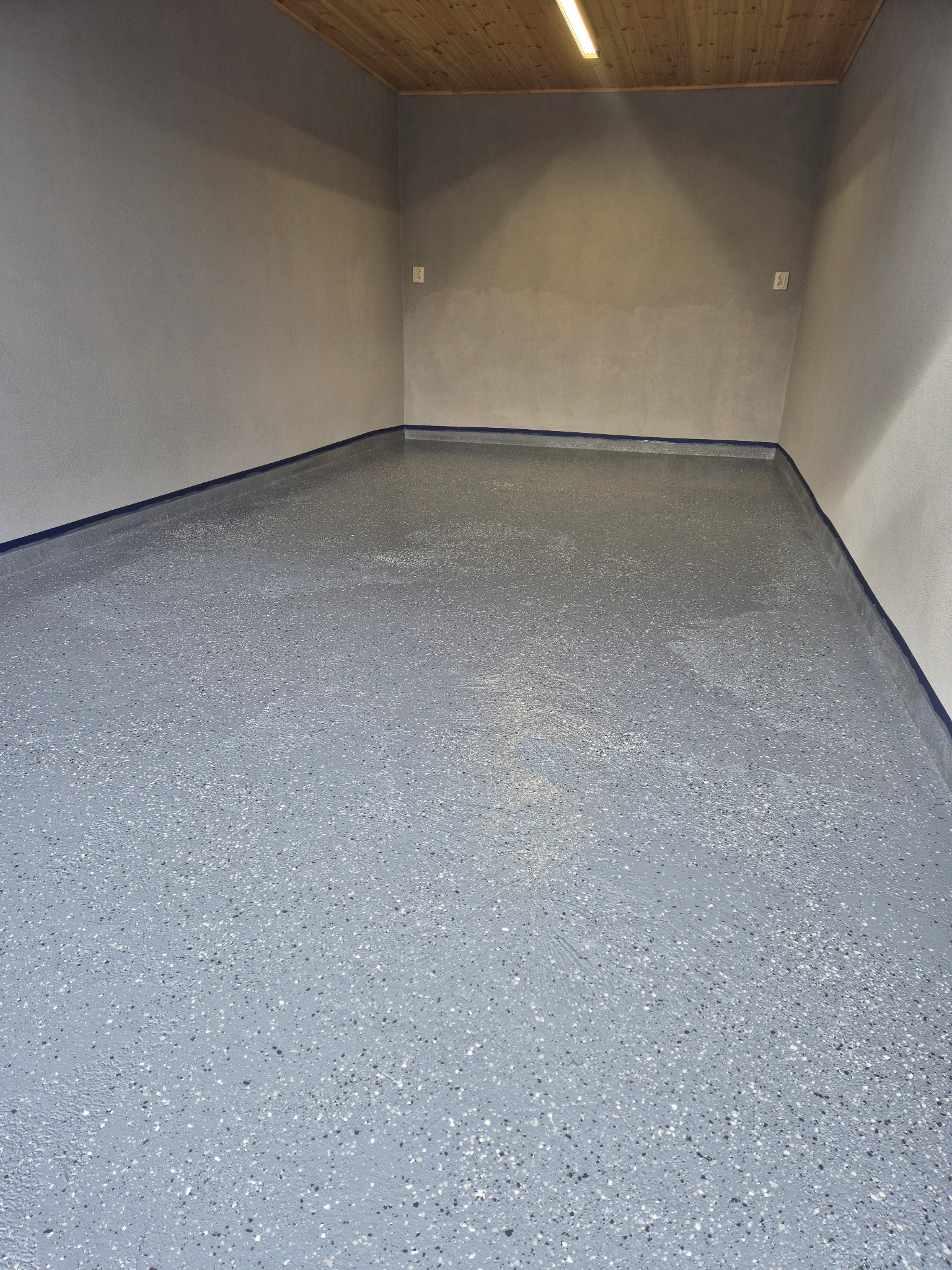Szara posadzka żywiczna z ciemnymi i jasnymi drobinkami, zabezpieczona niebieską taśmą malarską przy ścianach w pustym pomieszczeniu z drewnianym sufitem.