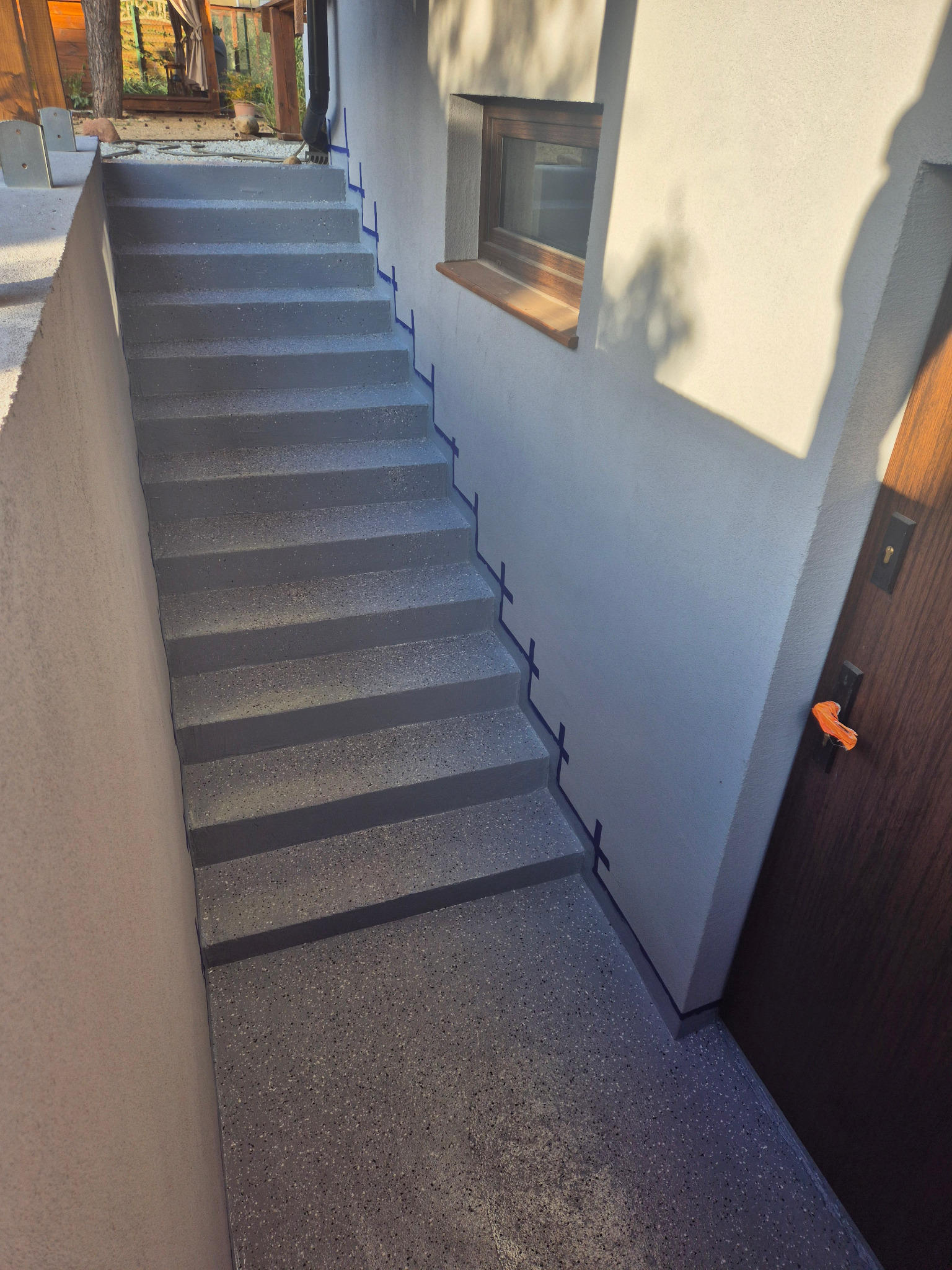 Zewnętrzne schody z szarego lastryko prowadzące do drzwi wejściowych, zabezpieczone niebieską taśmą malarską przy ścianie.
