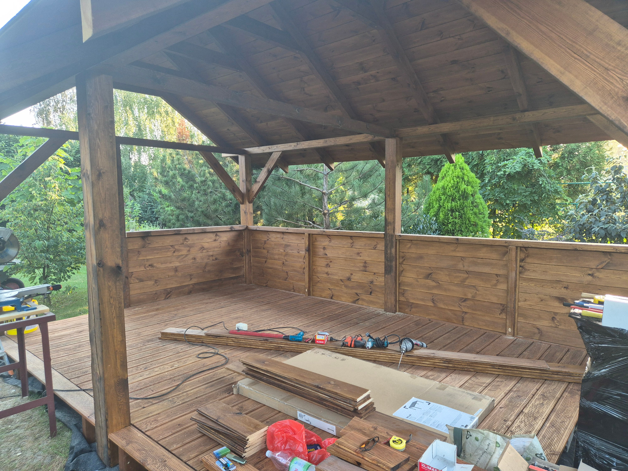 Drewniana altana w trakcie budowy: deski podłogowe, narzędzia, widok na zieleń w tle. Widoczne puszki z impregnatem do drewna.