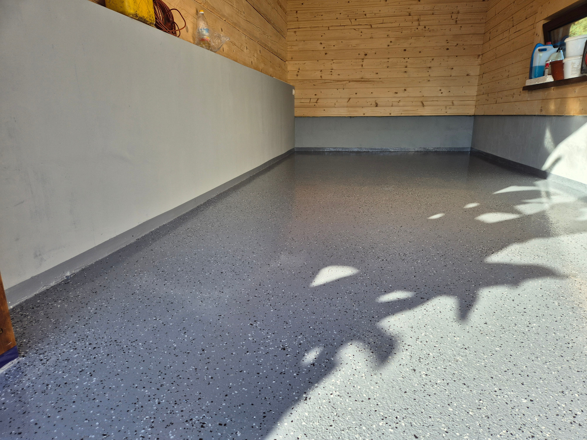 Świeżo wylana, błyszcząca podłoga żywiczna w garażu lub pomieszczeniu gospodarczym, z widocznymi drobnymi, kolorowymi wtrąceniami, szare ściany i drewniane wykończenie ścian na górze, cień rzucany...
