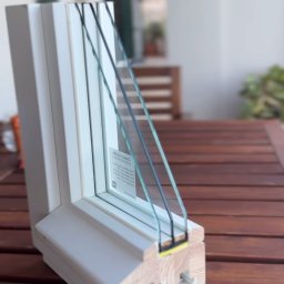 HLO Okna Fenster z polski - Przekrój ramy okiennej z trzema szybami, ukazujący szczegóły konstrukcji i materiały wykończeniowe na drewnianym stole.