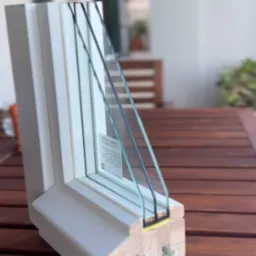 Przekrój ramy okiennej z trzema szybami, ukazujący szczegóły konstrukcji i materiały wykończeniowe na drewnianym stole.